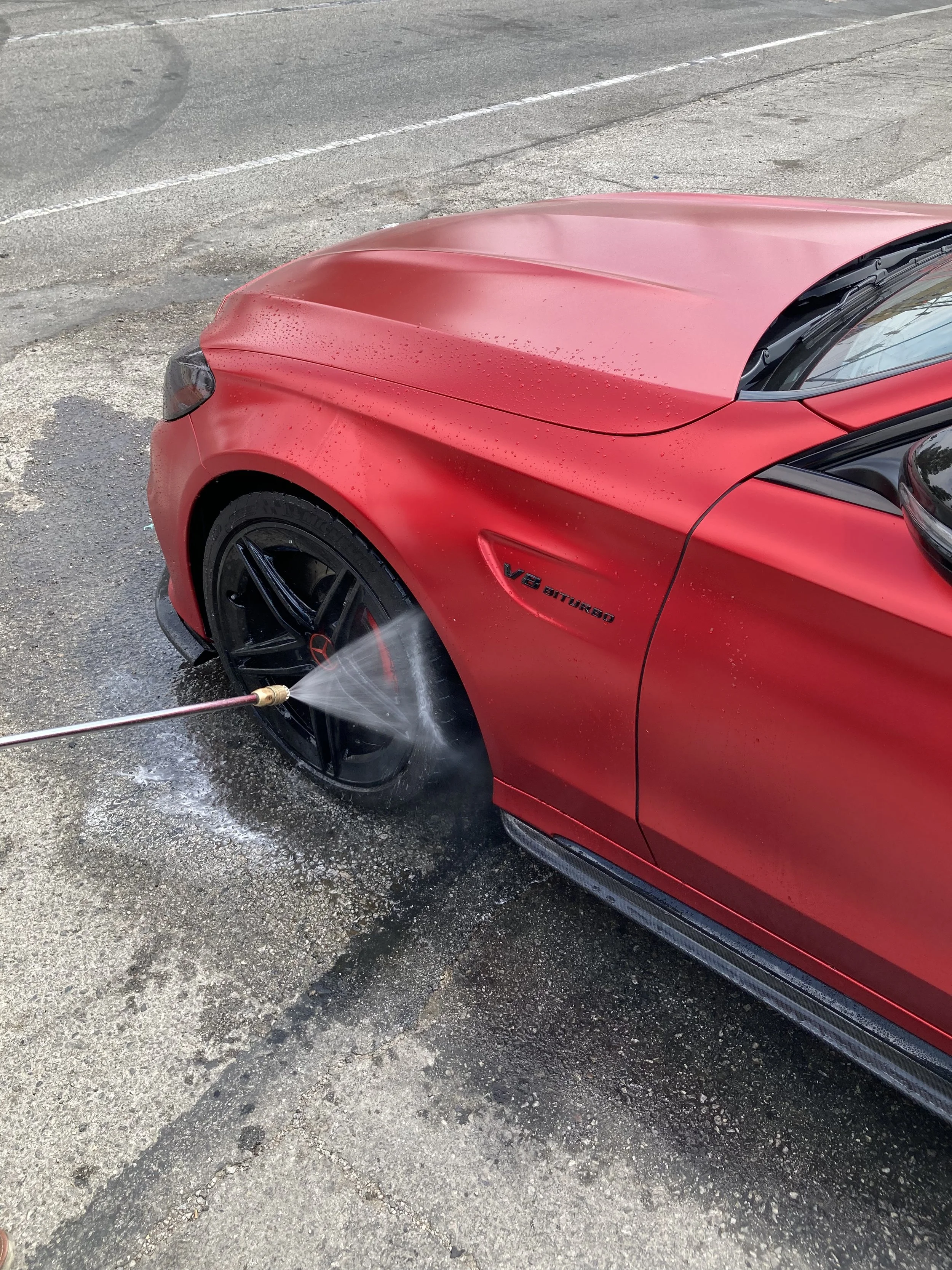 A red Mercedes-Benz car being washed with a pressure washer on its front left tire and fender.