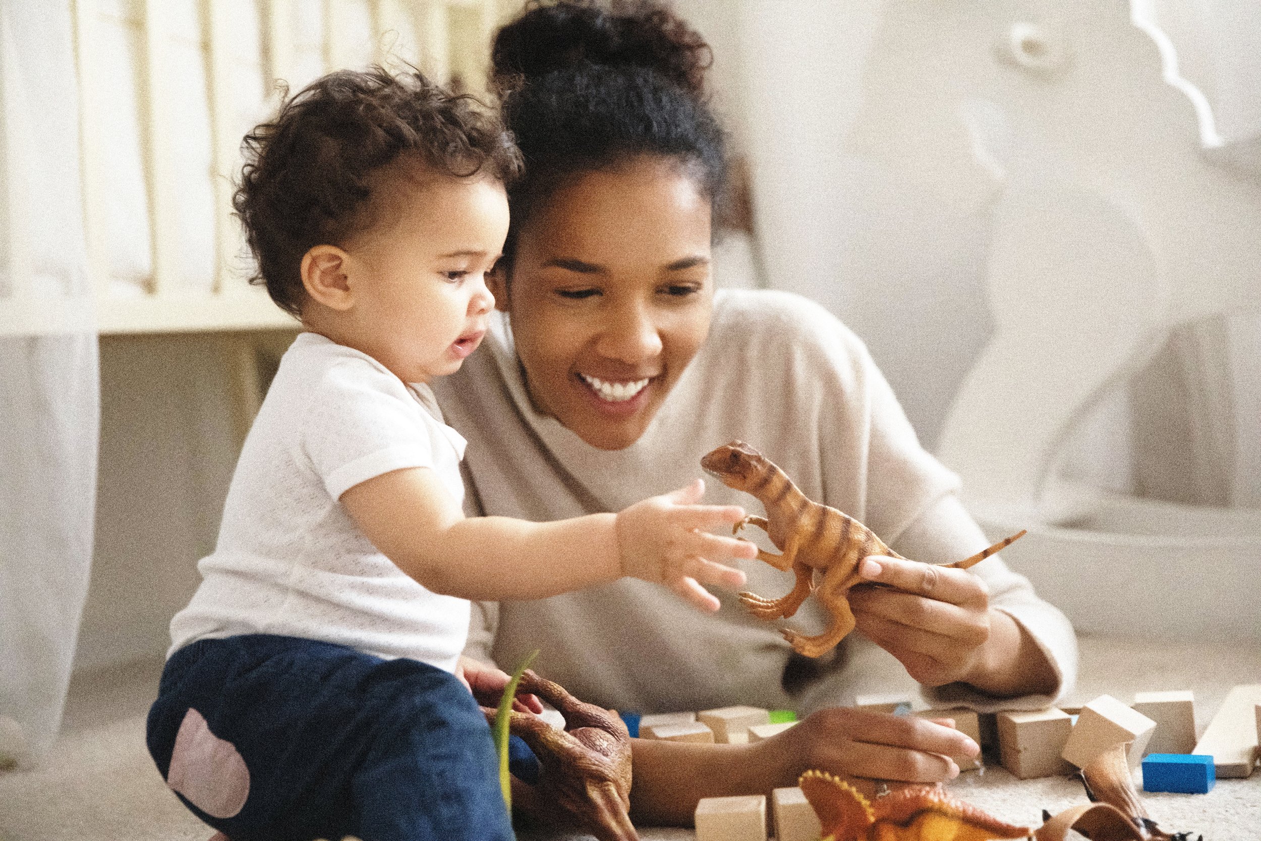A woman and a young child playing with toy dinosaurs on the floor in a cozy room.