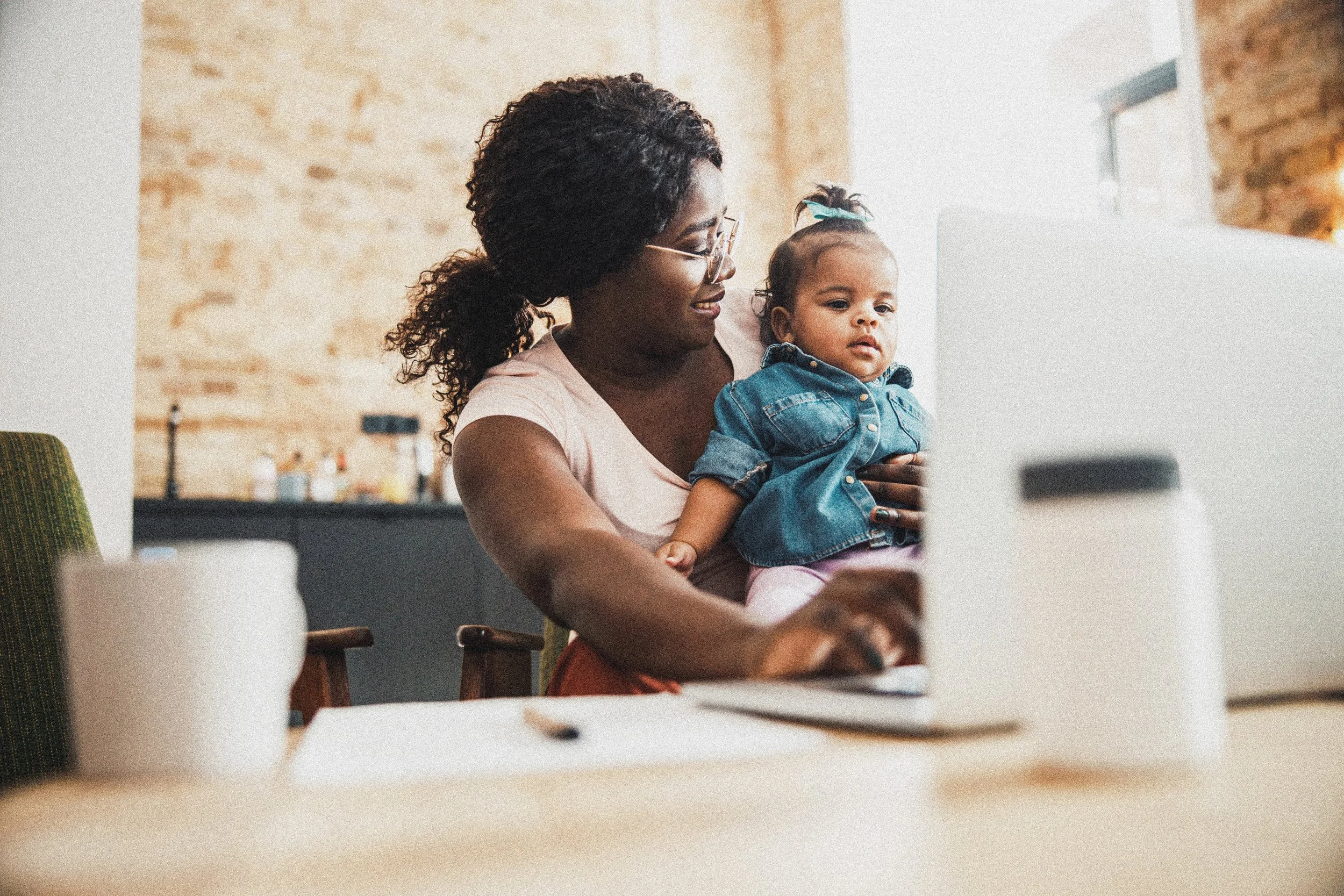 A woman with curly hair and glasses holding a young girl with a ponytail and denim jacket while working on a computer in a cozy, well-lit room with a brick wall in the background.