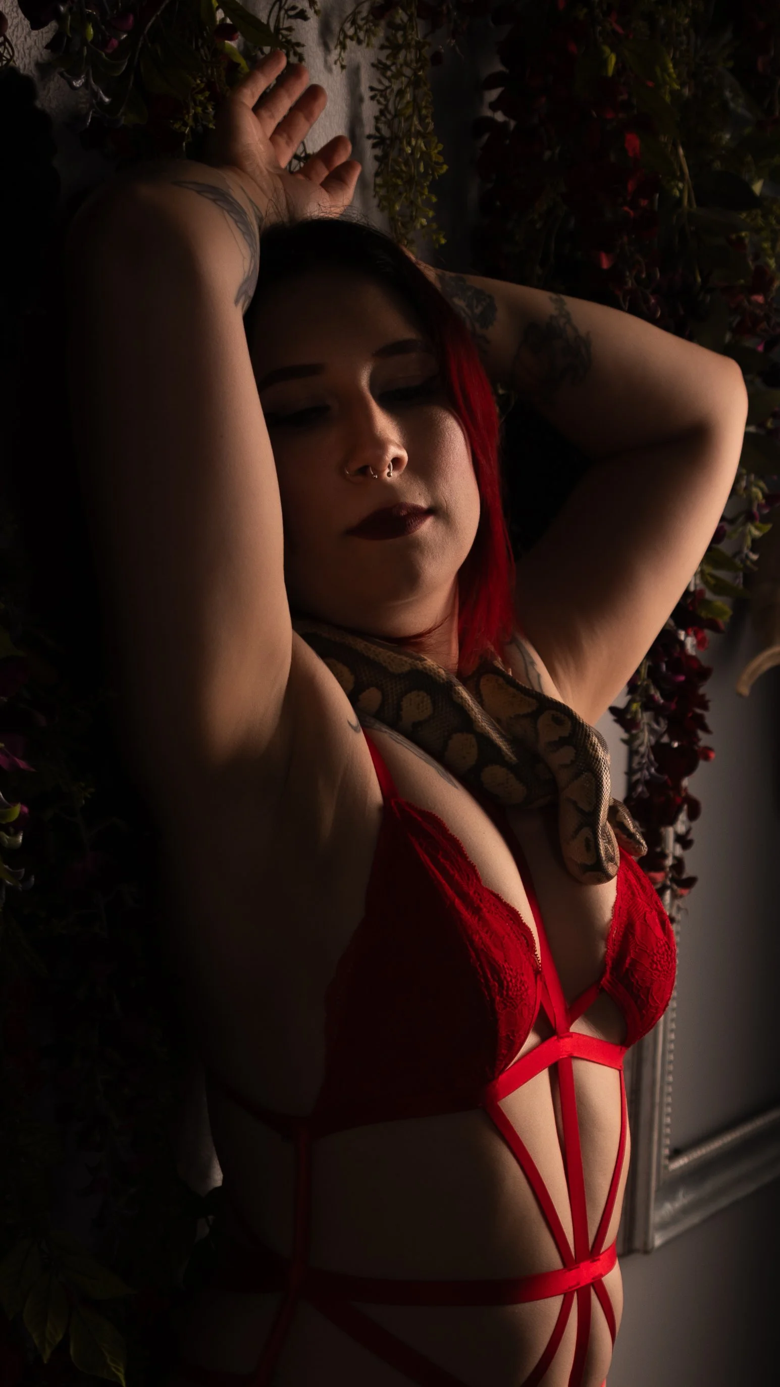A woman with red hair, tattoos, and dark makeup lying down with her eyes closed. She is wearing red lace lingerie and has a snake draped around her neck. She is surrounded by plants and flowers.
