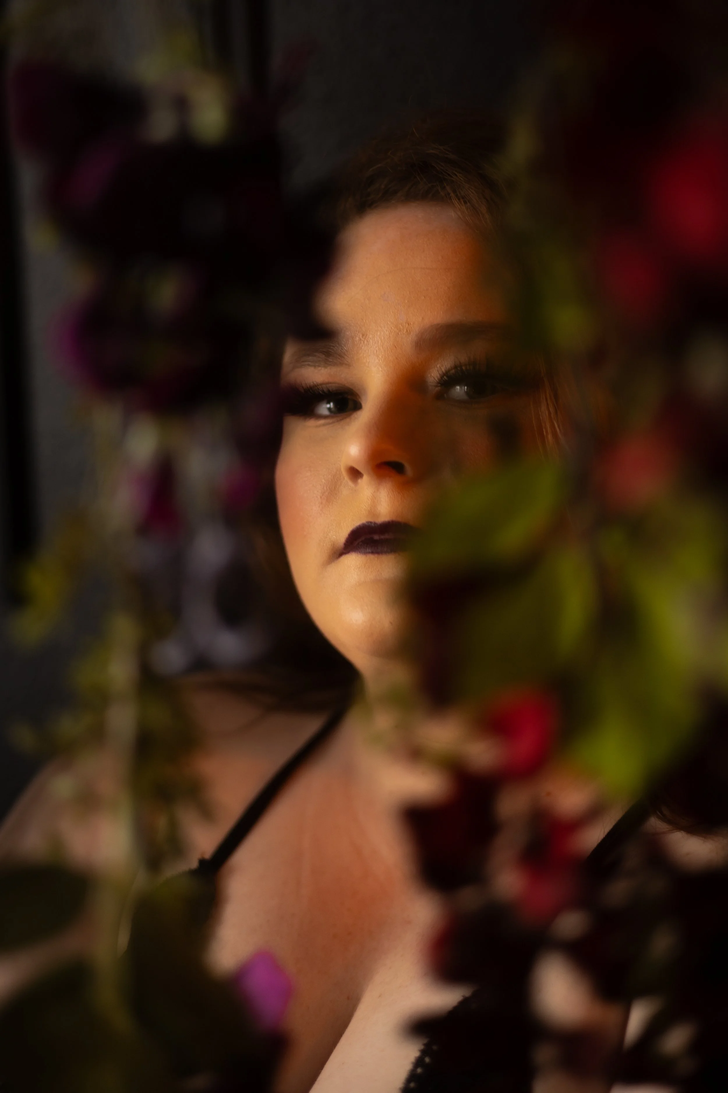 Close-up of a woman's face with dark makeup, partially obscured by out-of-focus colorful flowers in the foreground.