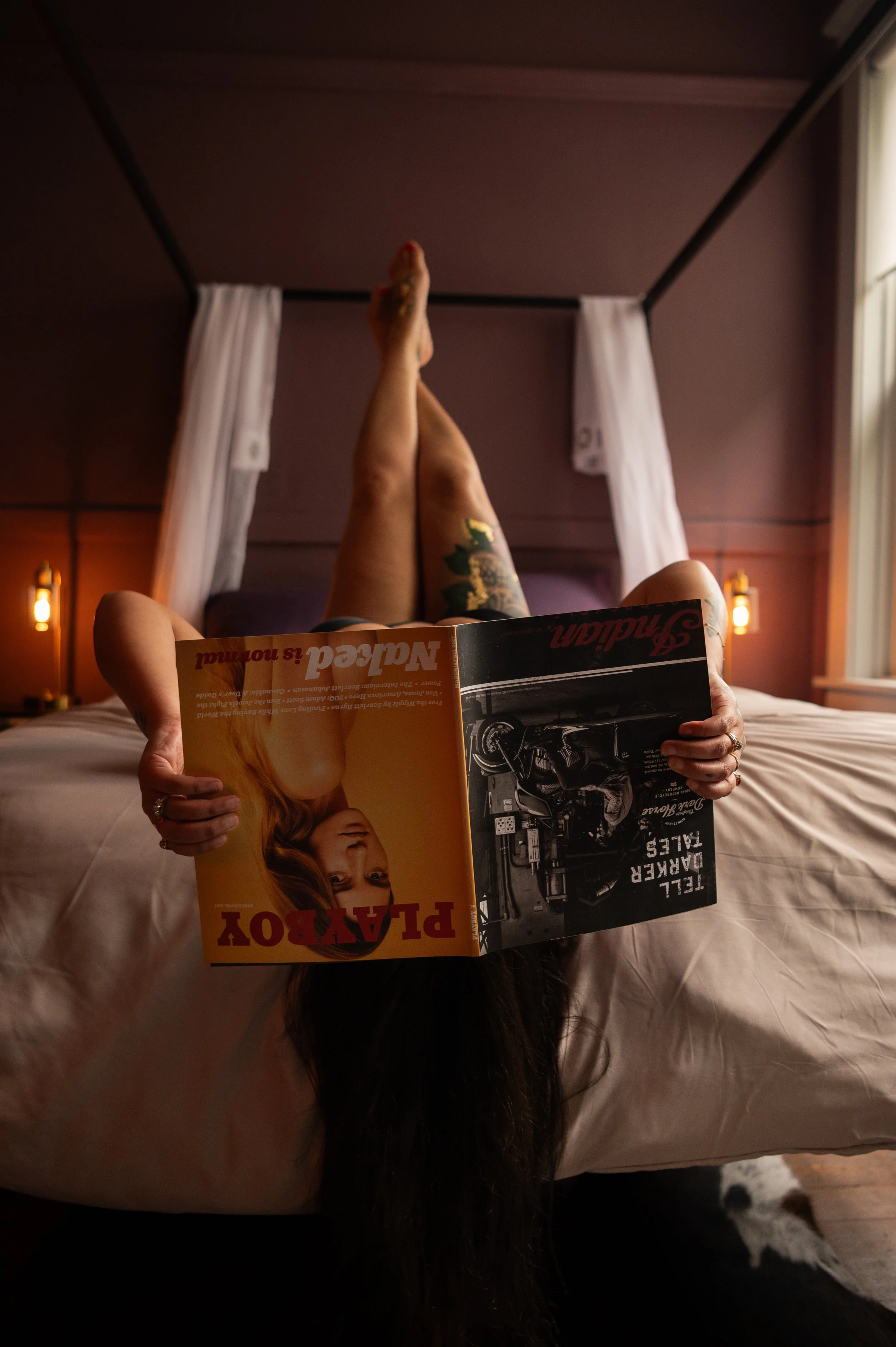 A person lying upside down on a bed, reading a magazine, with their head hanging off the edge and legs extended upward.