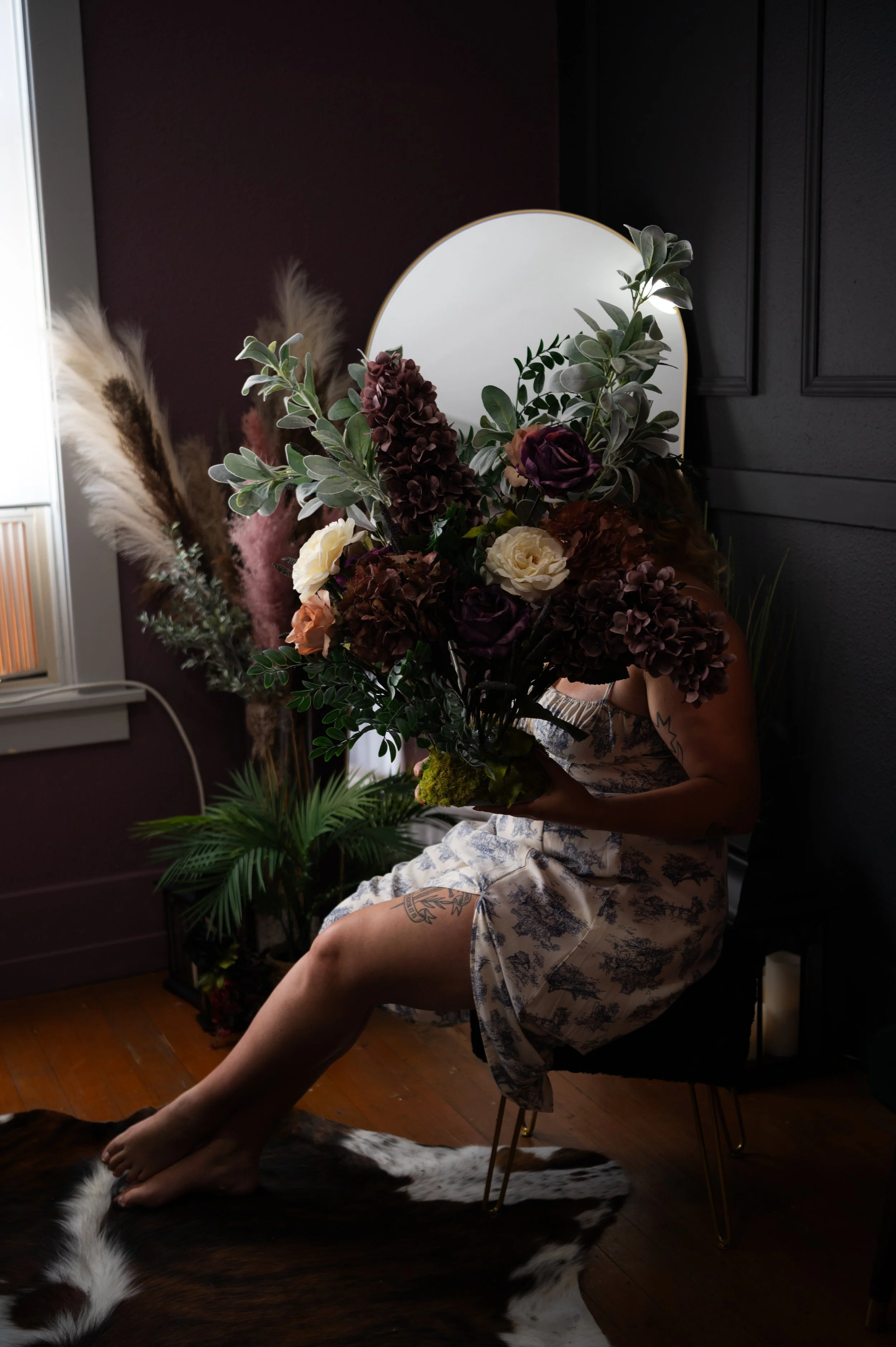 Person holding a large bouquet of flowers, sitting on a chair in a dimly lit room with dark walls and plants, with a cowhide rug on the floor.