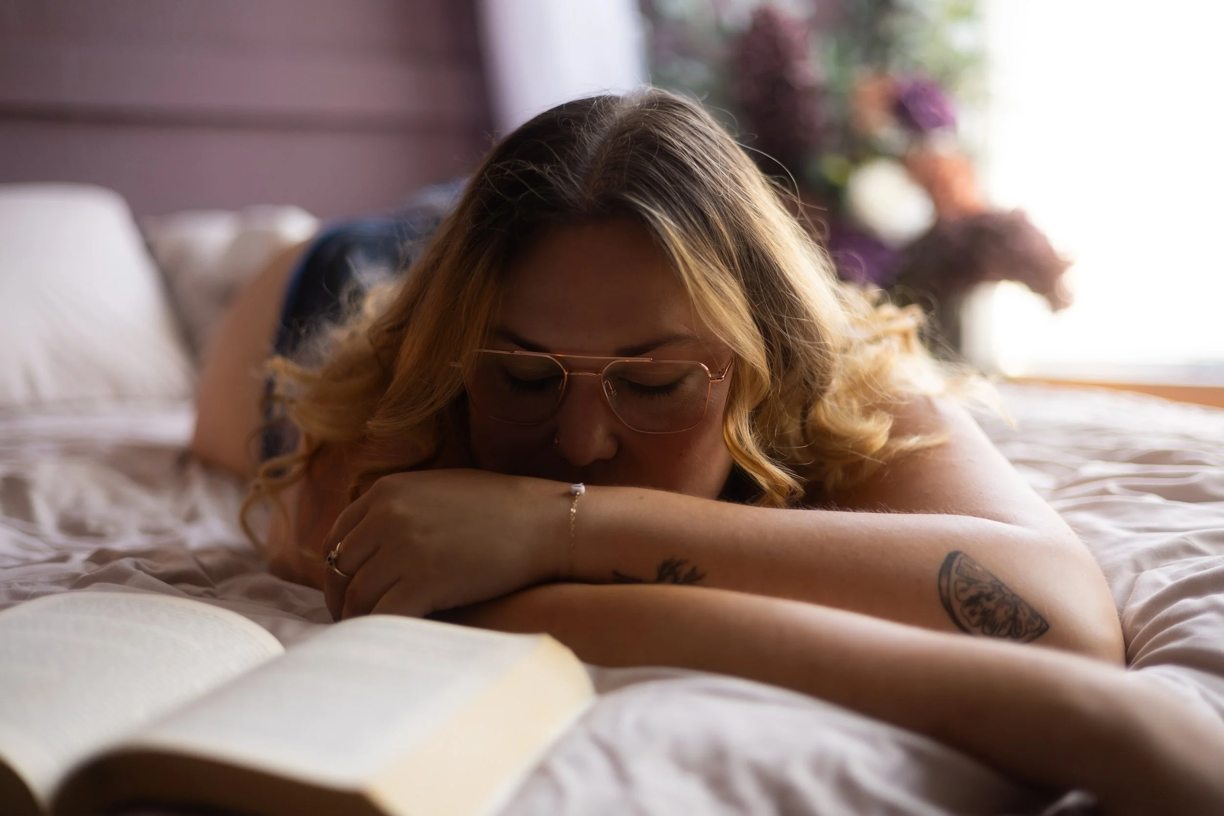 Woman with glasses lying on bed with arms crossed, reading a book, in a softly lit room with flowers in the background.
