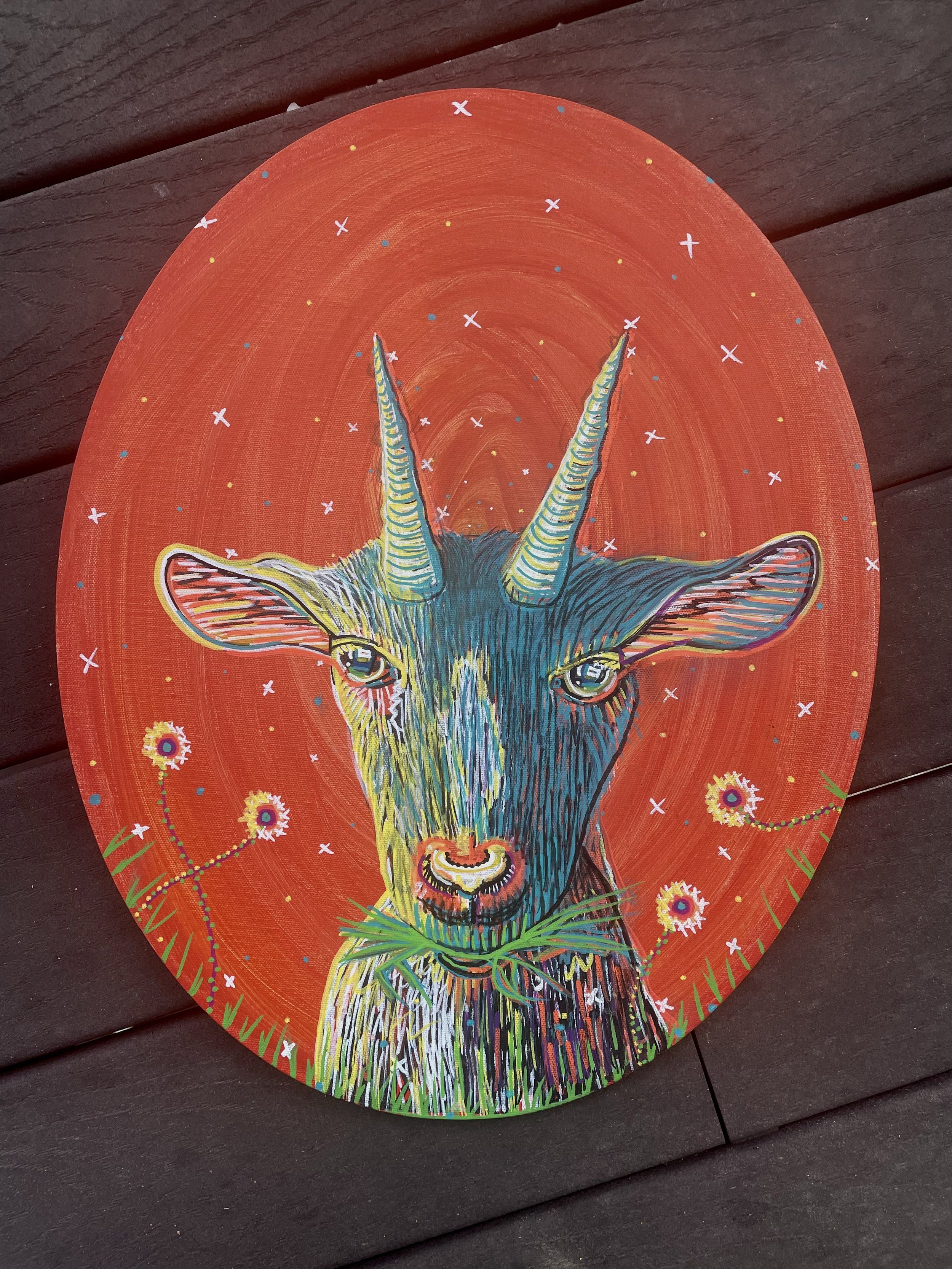 Colorful painting of a goat with striped horns and a tuft of grass in its mouth, set against an orange background with stars and dandelions.