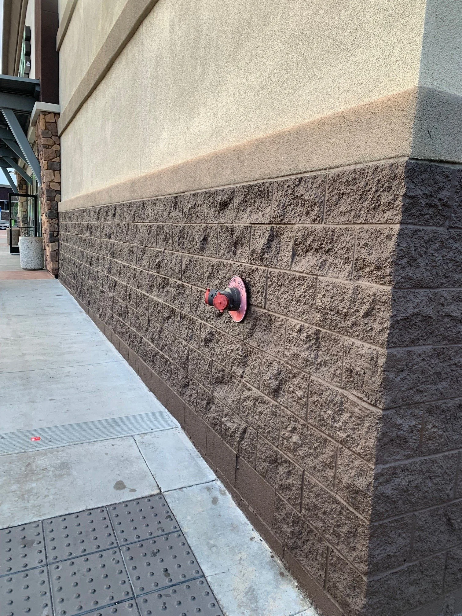 Exterior view of a building wall with a fire hydrant fitting on the brick section near the sidewalk.