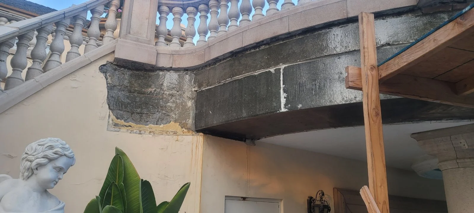 Close-up of a corner of a building with exterior repairs, showing a damaged stone edge, part of a decorative stone railing, a white bust sculpture, and some green plants.