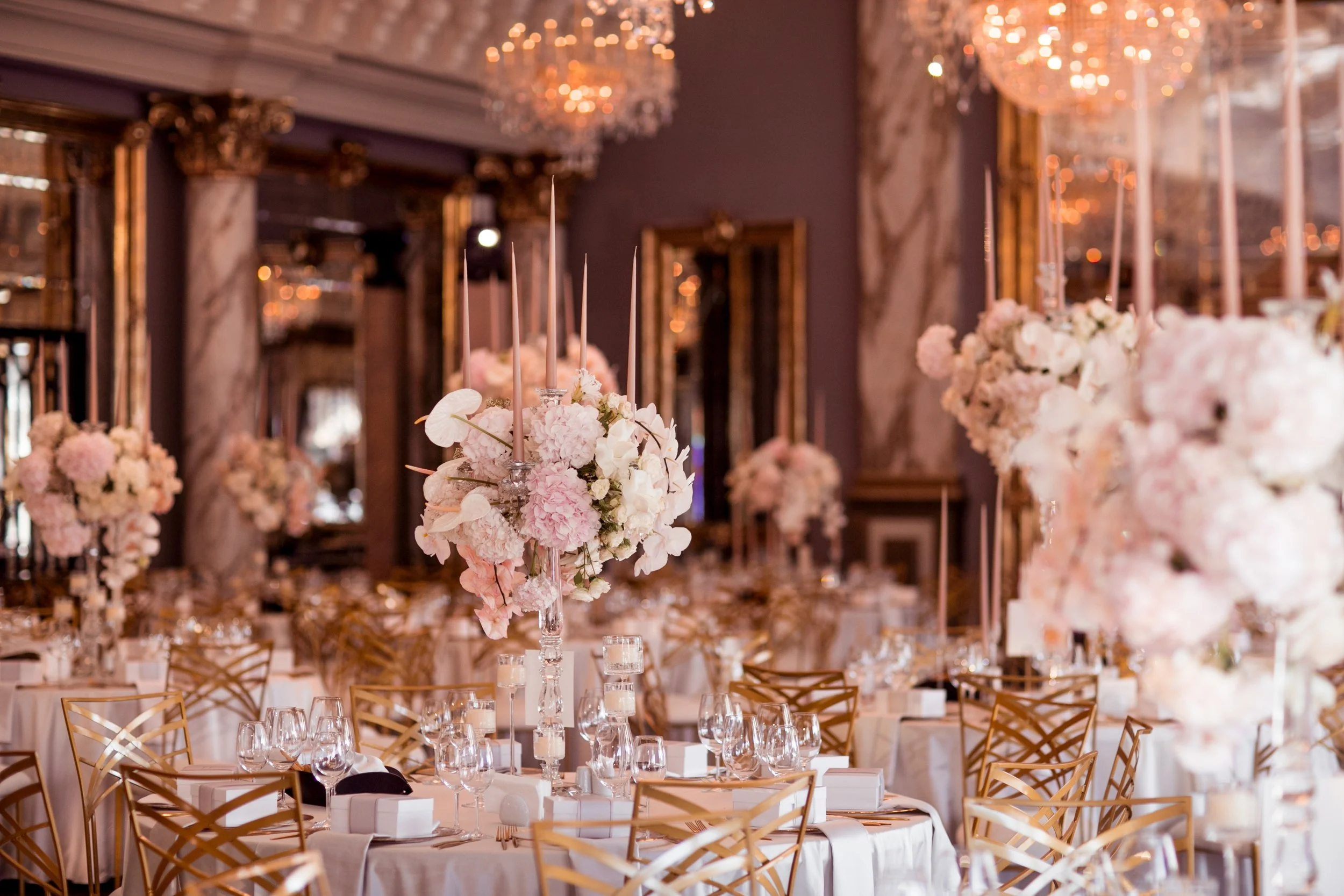 Elegant banquet hall decorated for a formal event with tall floral centerpieces, hanging chandeliers, and gold chairs.