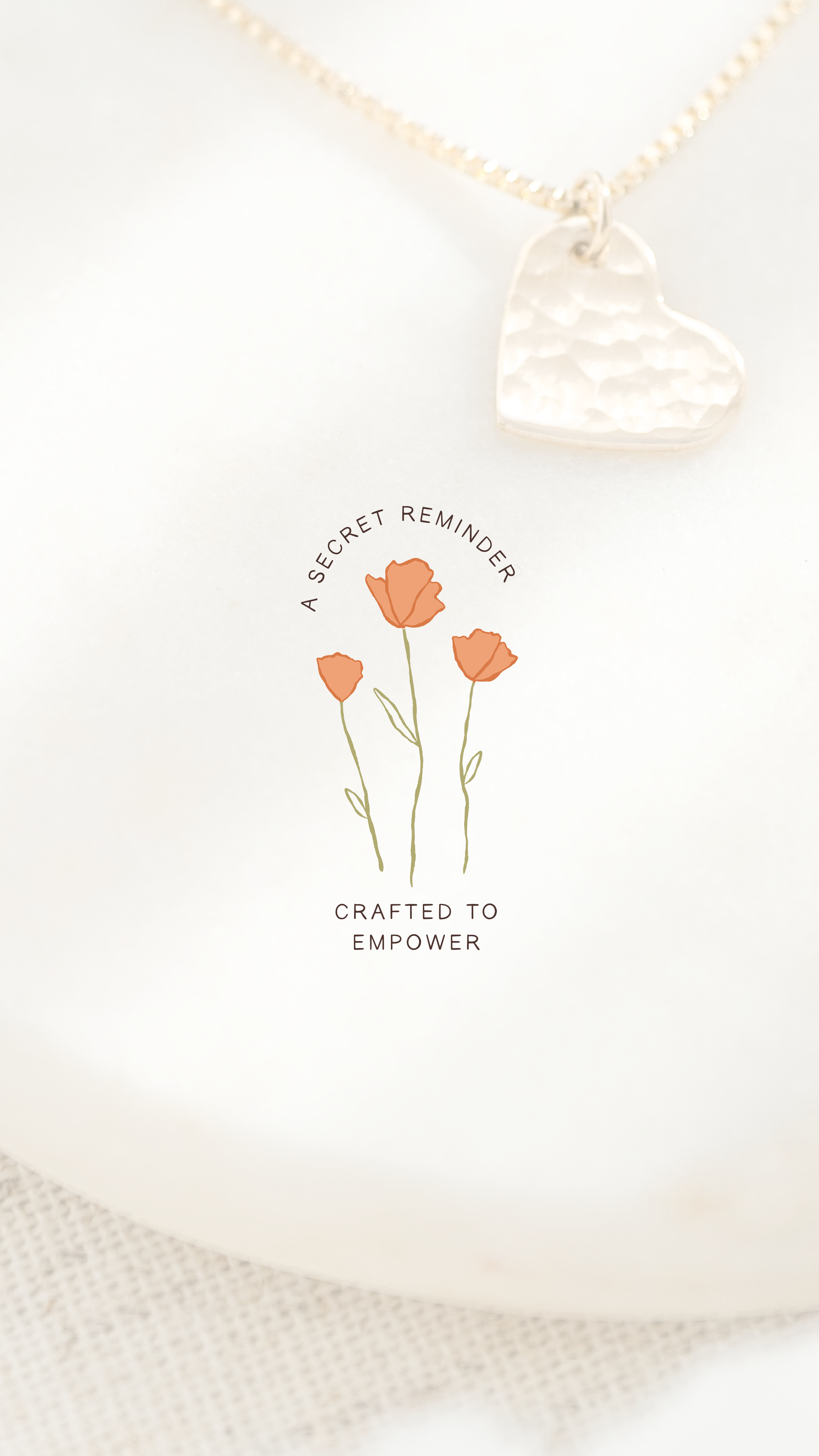 A silver necklace with a heart-shaped pendant rests on paper that has a drawing of three orange flowers and the text 'A SECRET REMINDER CRAFTED TO EMPOWER'.