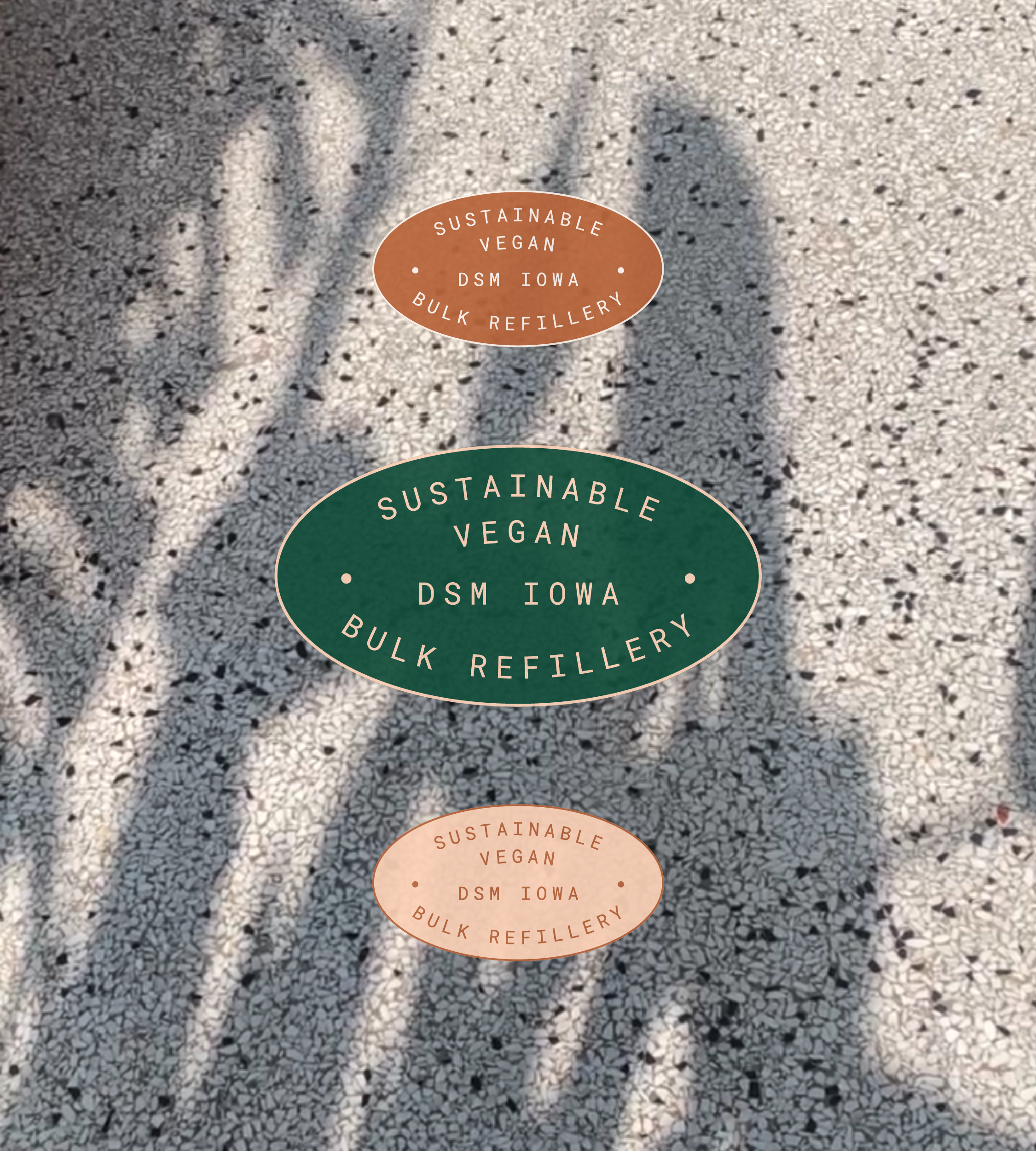 Three stickers on a textured surface with shadows of plants. The stickers read 'Sustainable Vegan DSM Iowa Bulk Refillery,' with one sticker larger and green, and two smaller, peach-colored stickers.