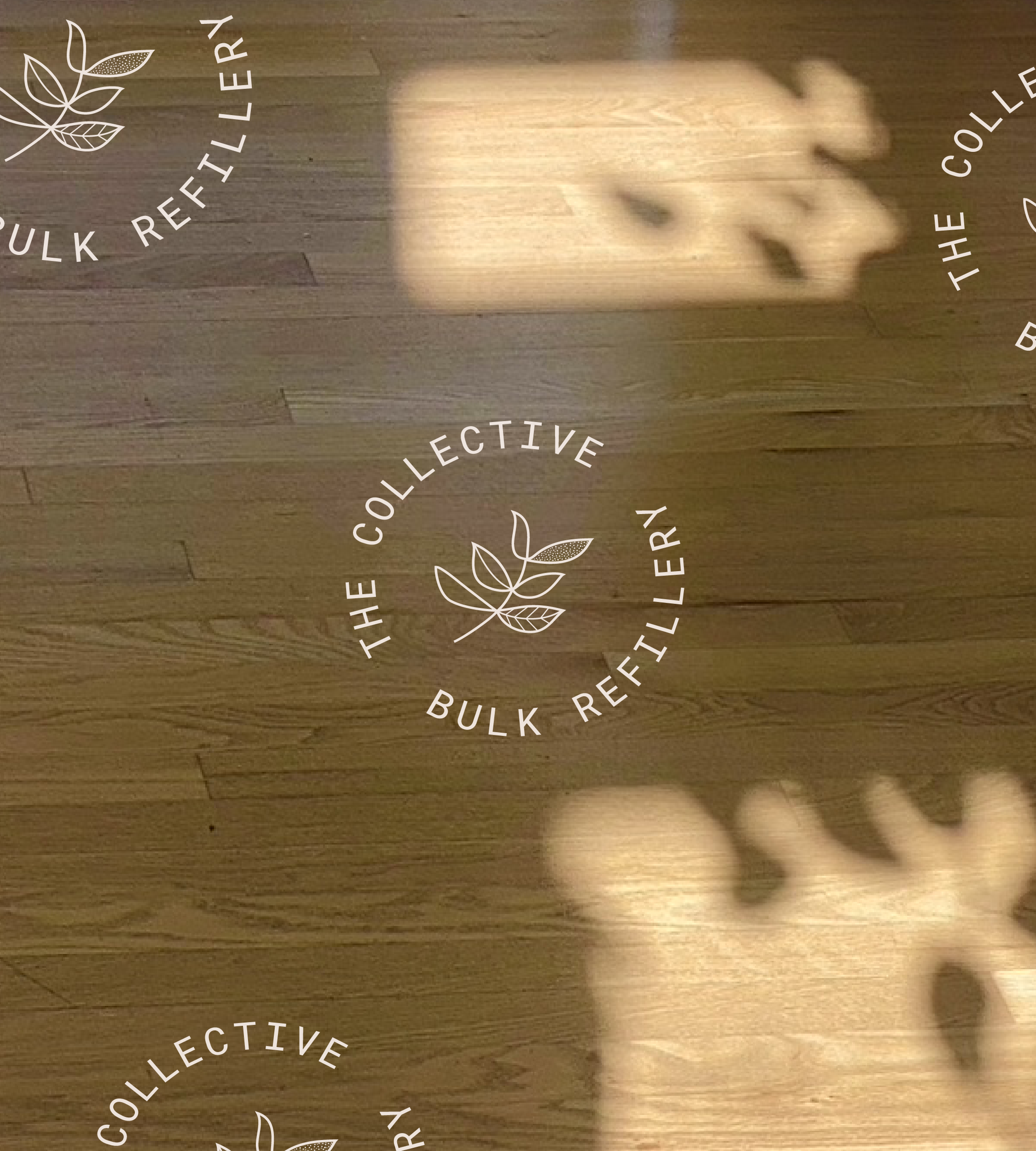 Wooden floor with shadows of large leaves, overlaid with white circular logo reading "The Collective Bulk Refillery" around a minimalist leaf illustration.