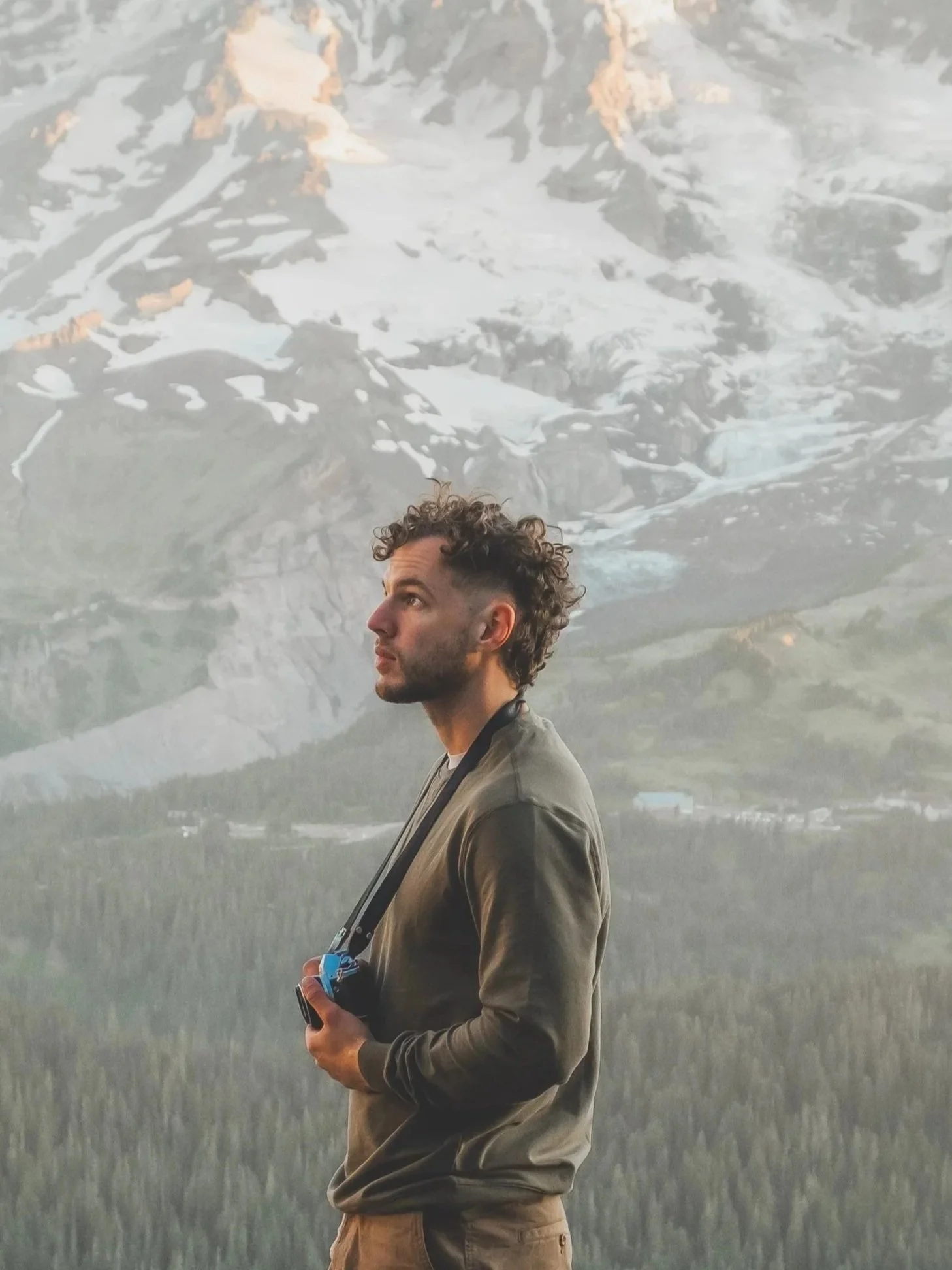 A man with curly hair holding a camera, standing outdoors with snow-capped mountains and forest in the background.