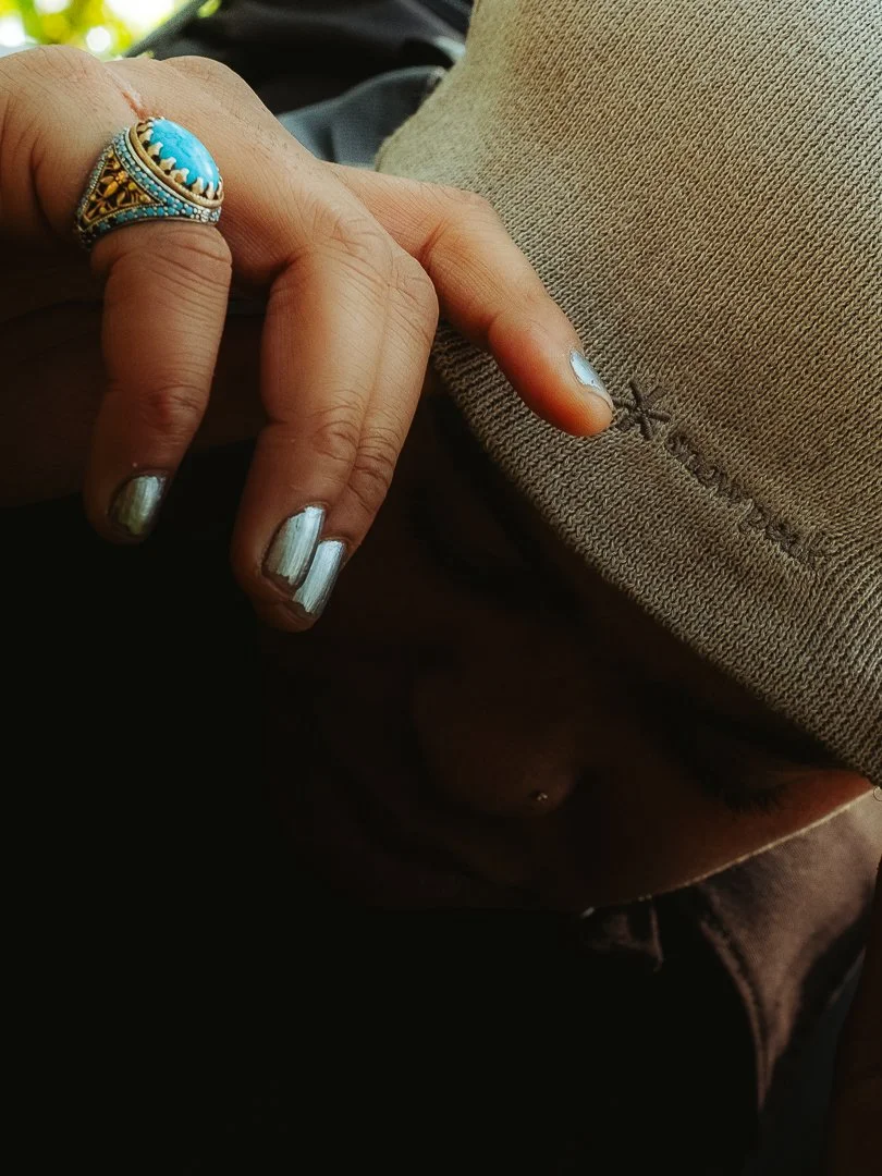 Close-up of a person's hand with metallic silver painted nails, wearing a turquoise and gold ring, touching a beige fabric with the word 'Xavier' embroidered on it.