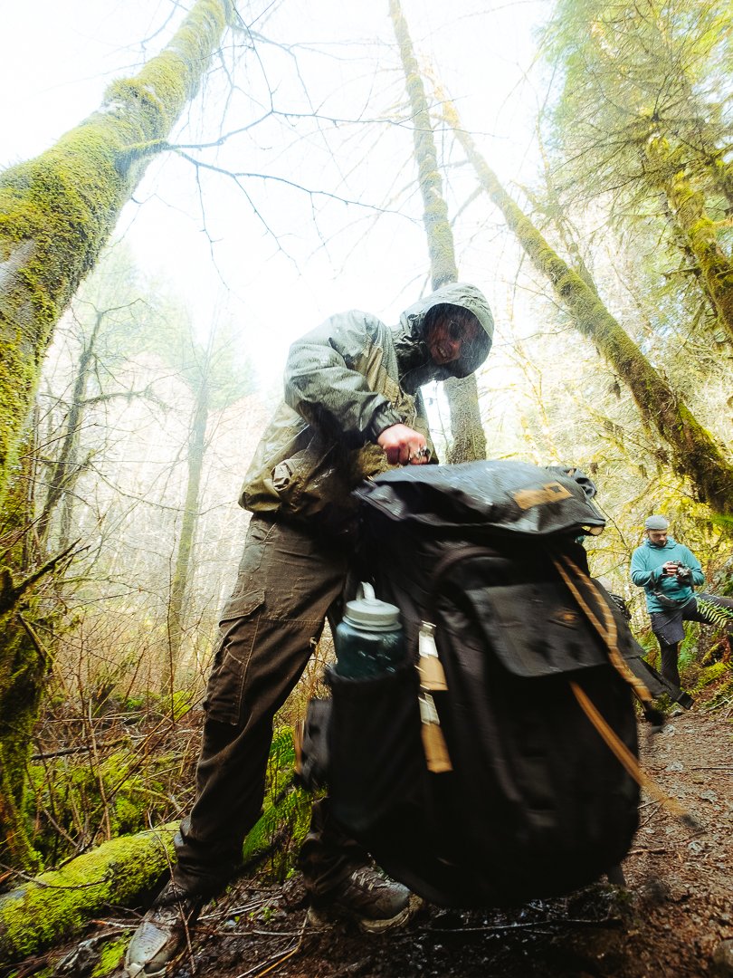 A person dressed in outdoor gear packing a large black backpack on a forest trail during foggy weather, with moss-covered trees and another person in the background looking at a device.