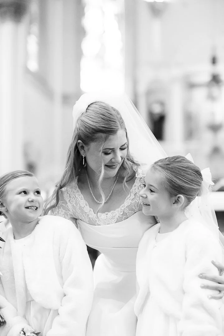 A bride in a wedding dress with a veil is smiling and leaning toward two young girls dressed in white, inside a church or cathedral.