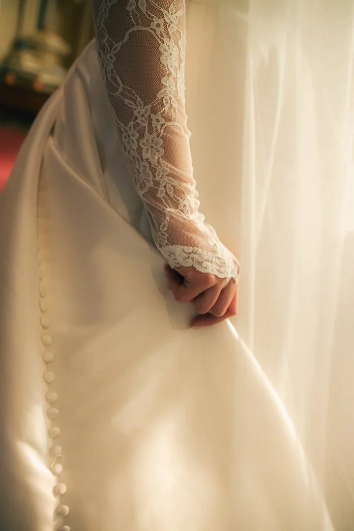 Close-up of a person wearing a wedding or bridal gown, focusing on their hand holding the fabric. The gown features lace details on the sleeve and satin buttons along the back.