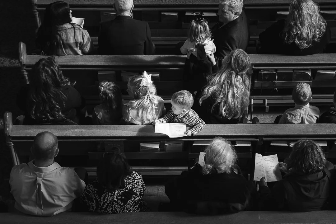 Black and white photo of people sitting in pews inside a church, facing the altar and engaged in a service or event, including children and adults.