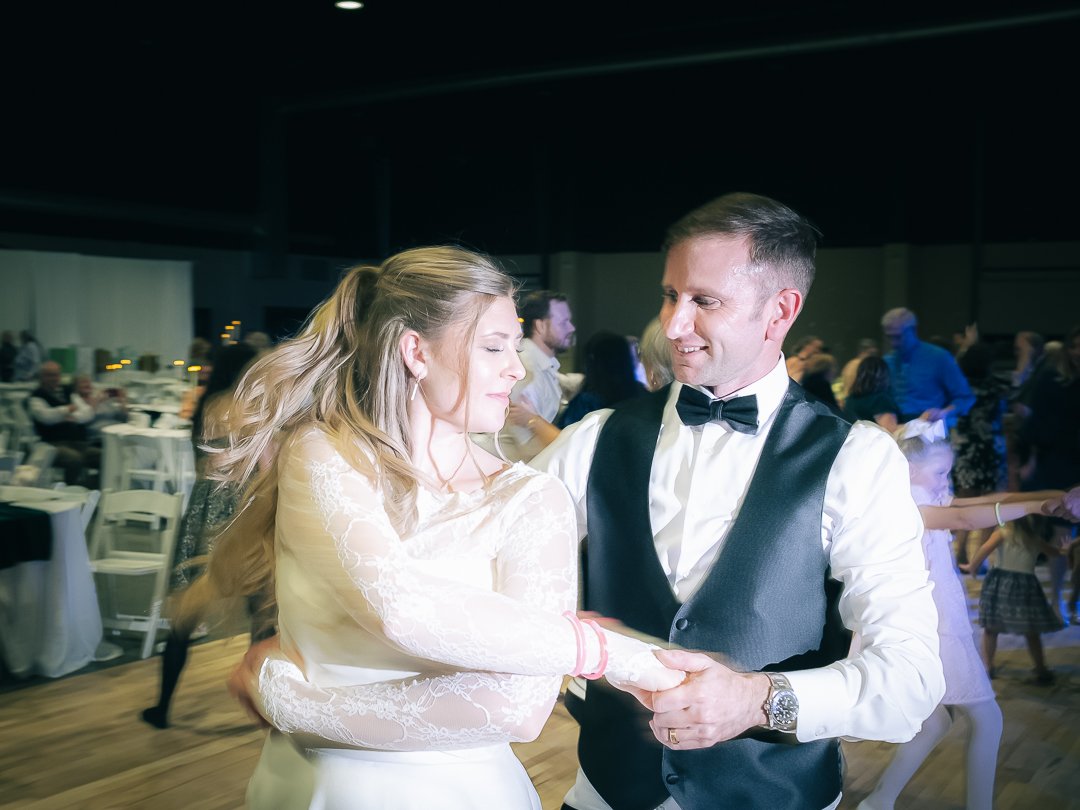 A couple dancing at a wedding reception, with the woman in a white dress and the man in a tuxedo, surrounded by guests.