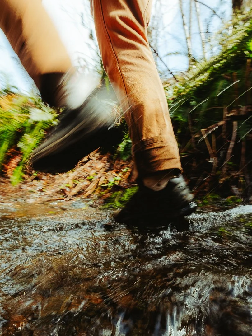 Person walking through a shallow creek, wearing brown pants and black shoes, with blurred motion in a forested area.