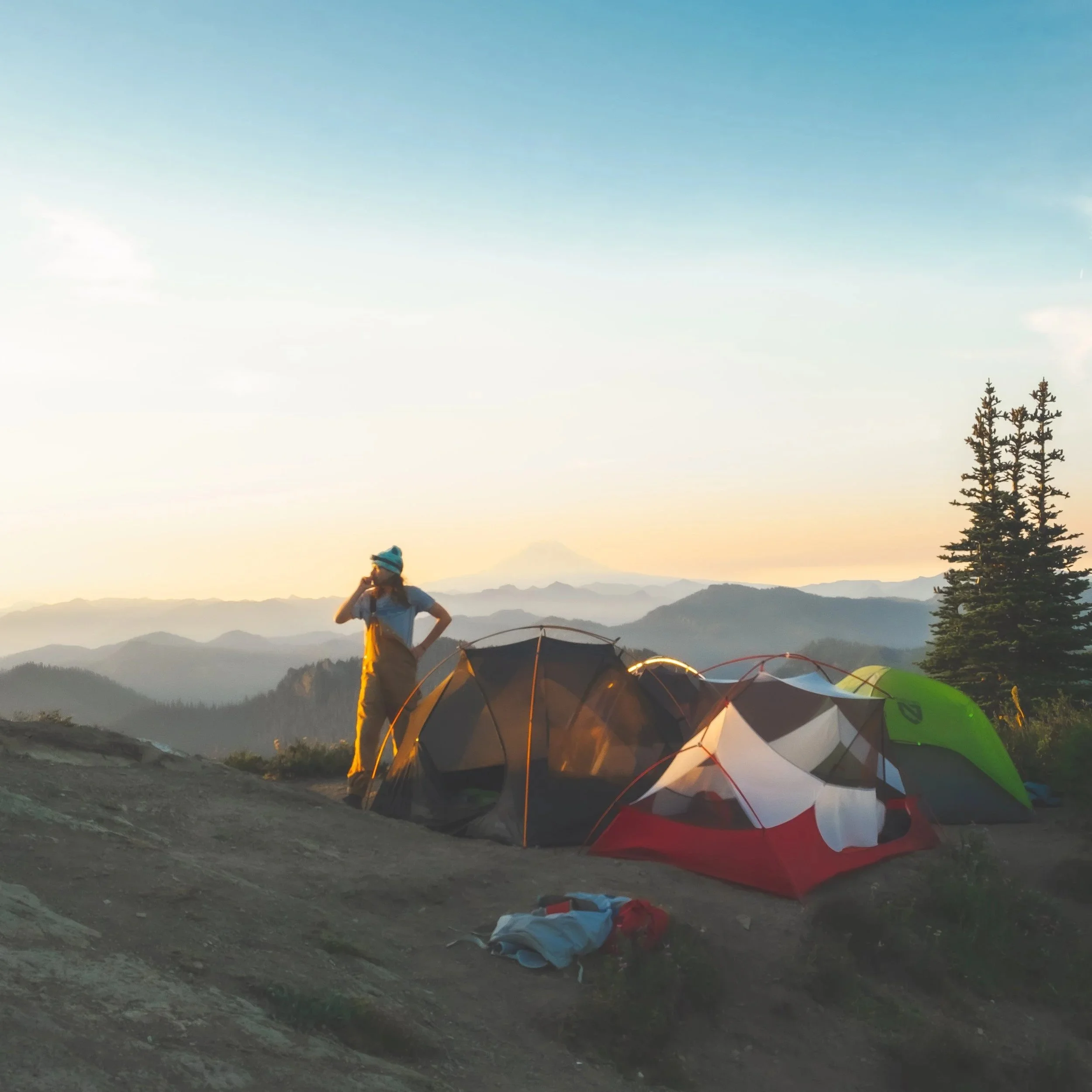 A woman standing near tents on a grassy hillside at sunrise with mountain ranges in the distance.
