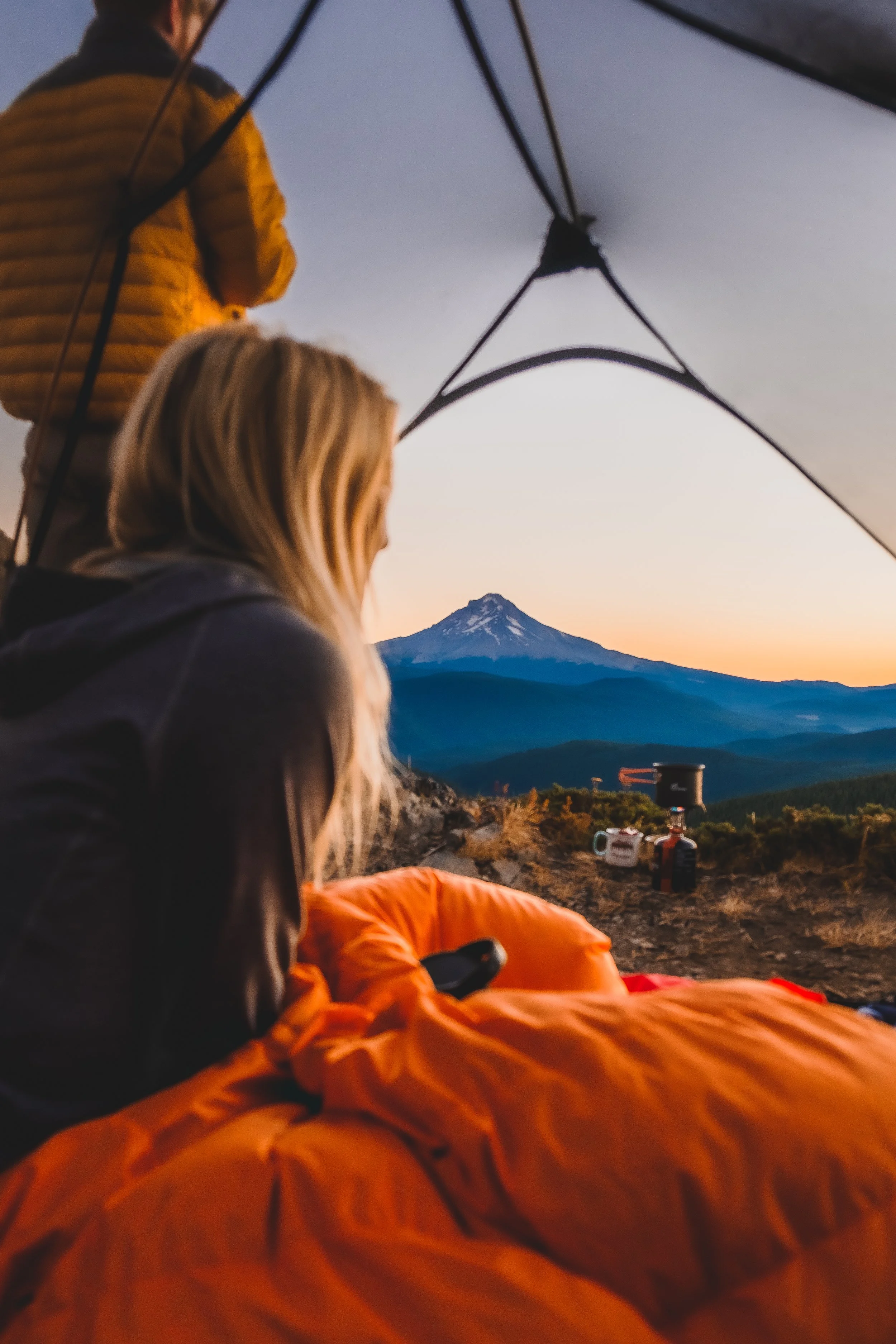 People camping inside a tent with a view of snow-capped Mount Hood at sunset.