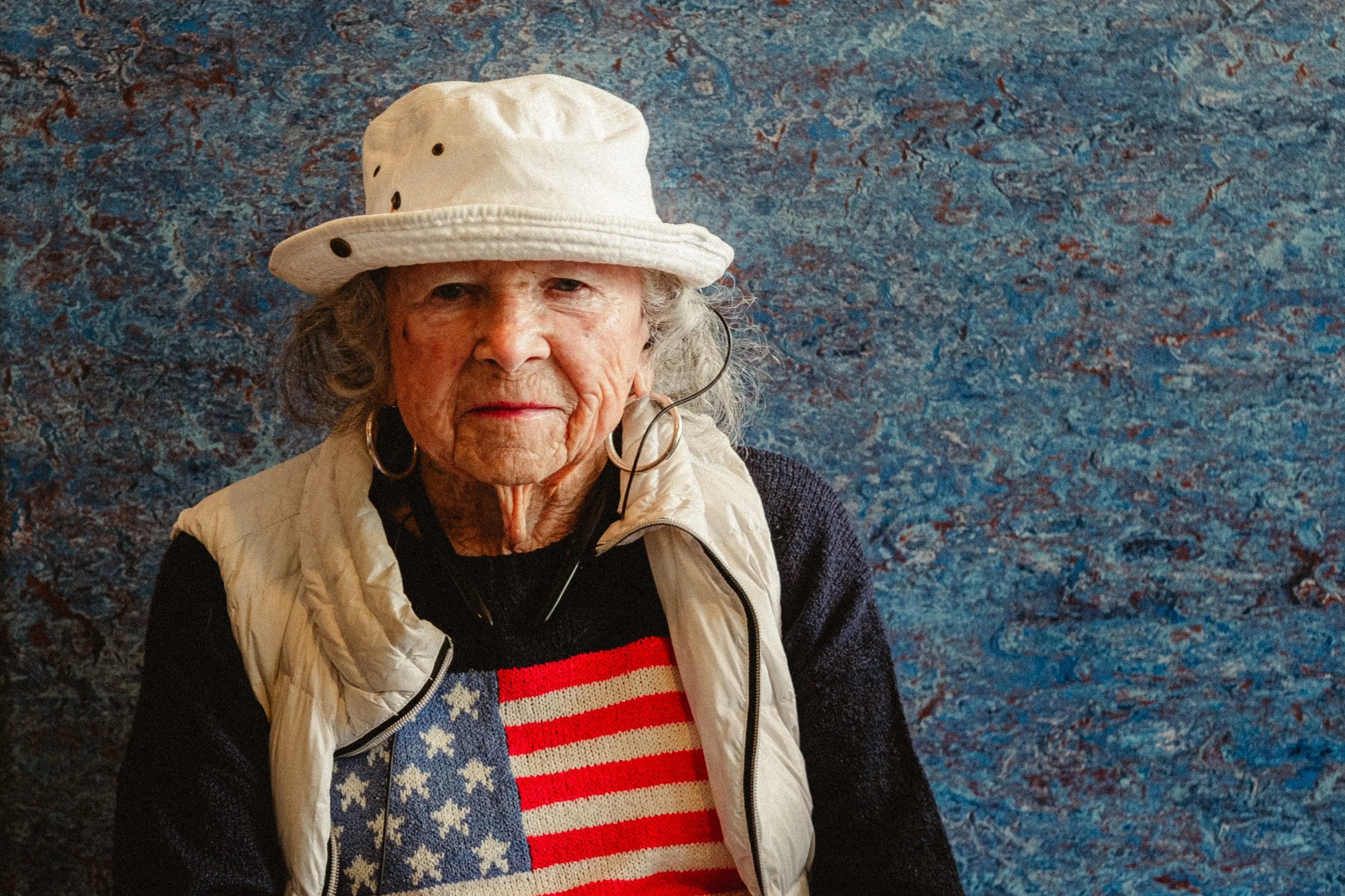 An elderly woman with gray hair and large earrings wearing a white hat, standing against a blue patterned background. She is dressed in a sweater with the American flag design and a white vest.