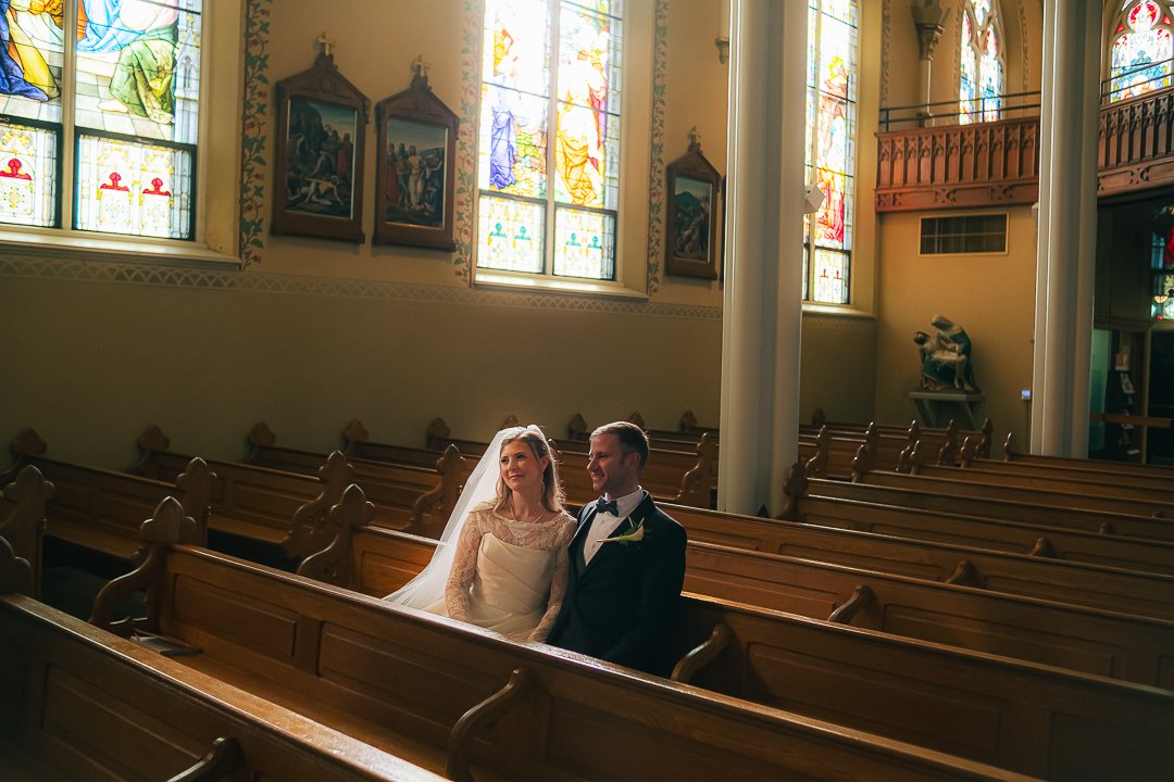 A bride and groom sitting in a church pew, with stained glass windows behind them.