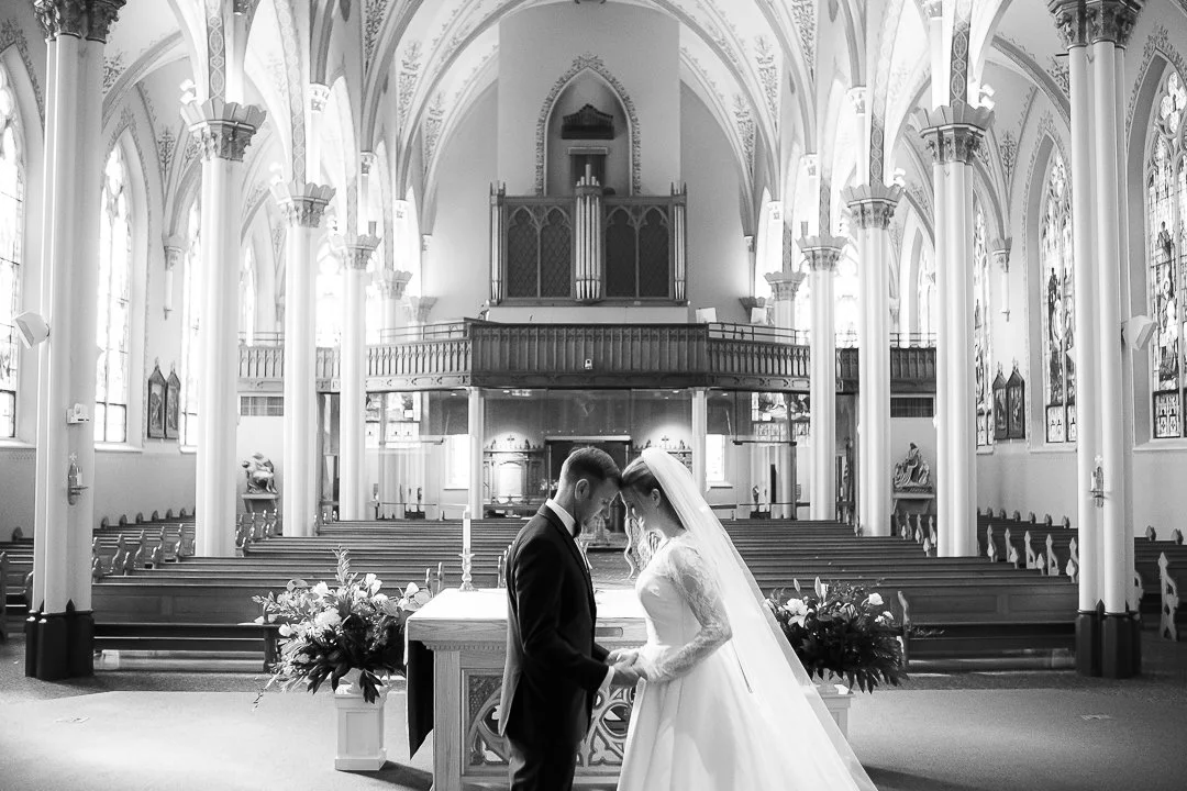 A bride and groom holding hands in a church during their wedding ceremony, with church pews and stained glass windows in the background.