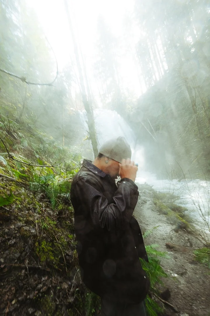 Person in a gray beanie and dark jacket standing on a damp forest path near a waterfall, holding sunglasses up to their face.
