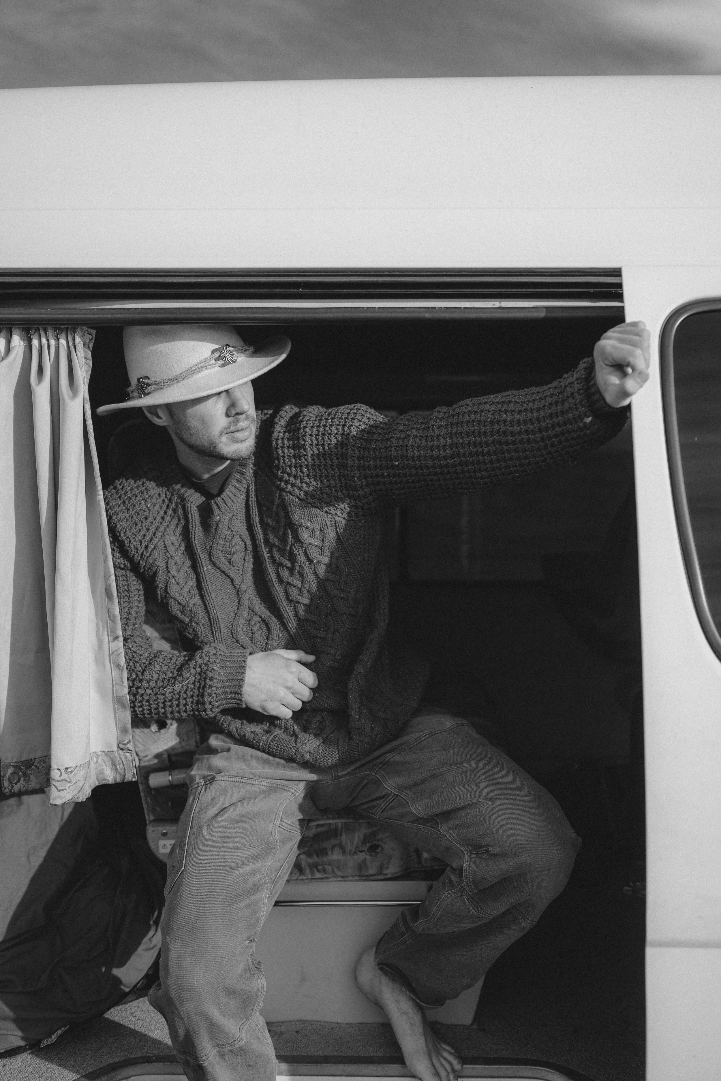 A man wearing a wide-brimmed hat and a knitted sweater leans out of a camper van, with his left arm extended and fist clenched.