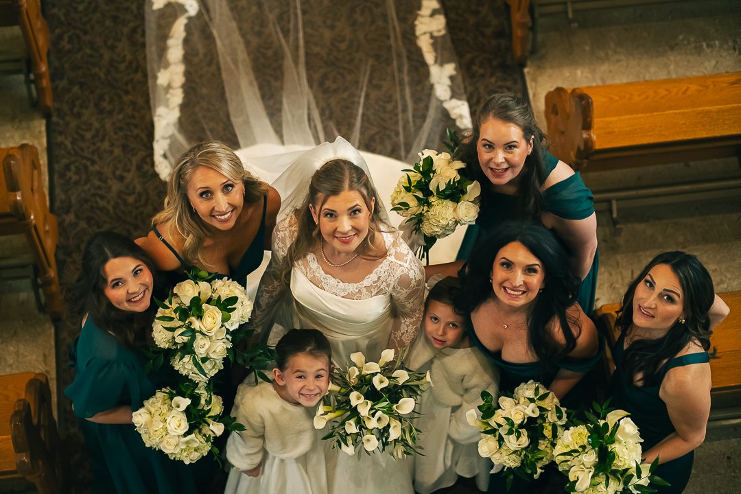 A group of women and children in wedding attire, gathered at a church, looking up at the camera and smiling. The women hold bouquets of white flowers.