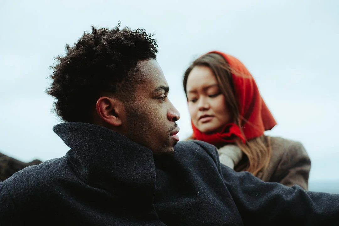 A man and woman outdoors, with the man in the foreground and woman in the background, both dressed warmly. The man has curly hair and a serious expression, looking straight ahead. The woman has long hair and a red headscarf, looking at the man with a