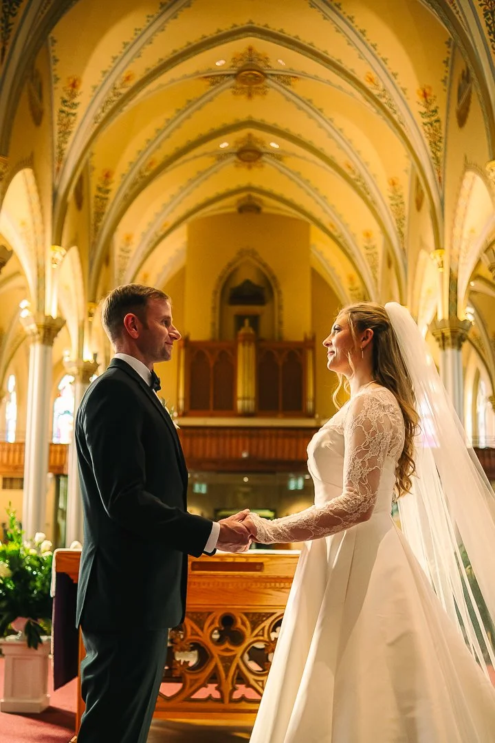 A bride and groom holding hands and looking at each other inside a church with ornate architecture and stained glass windows.