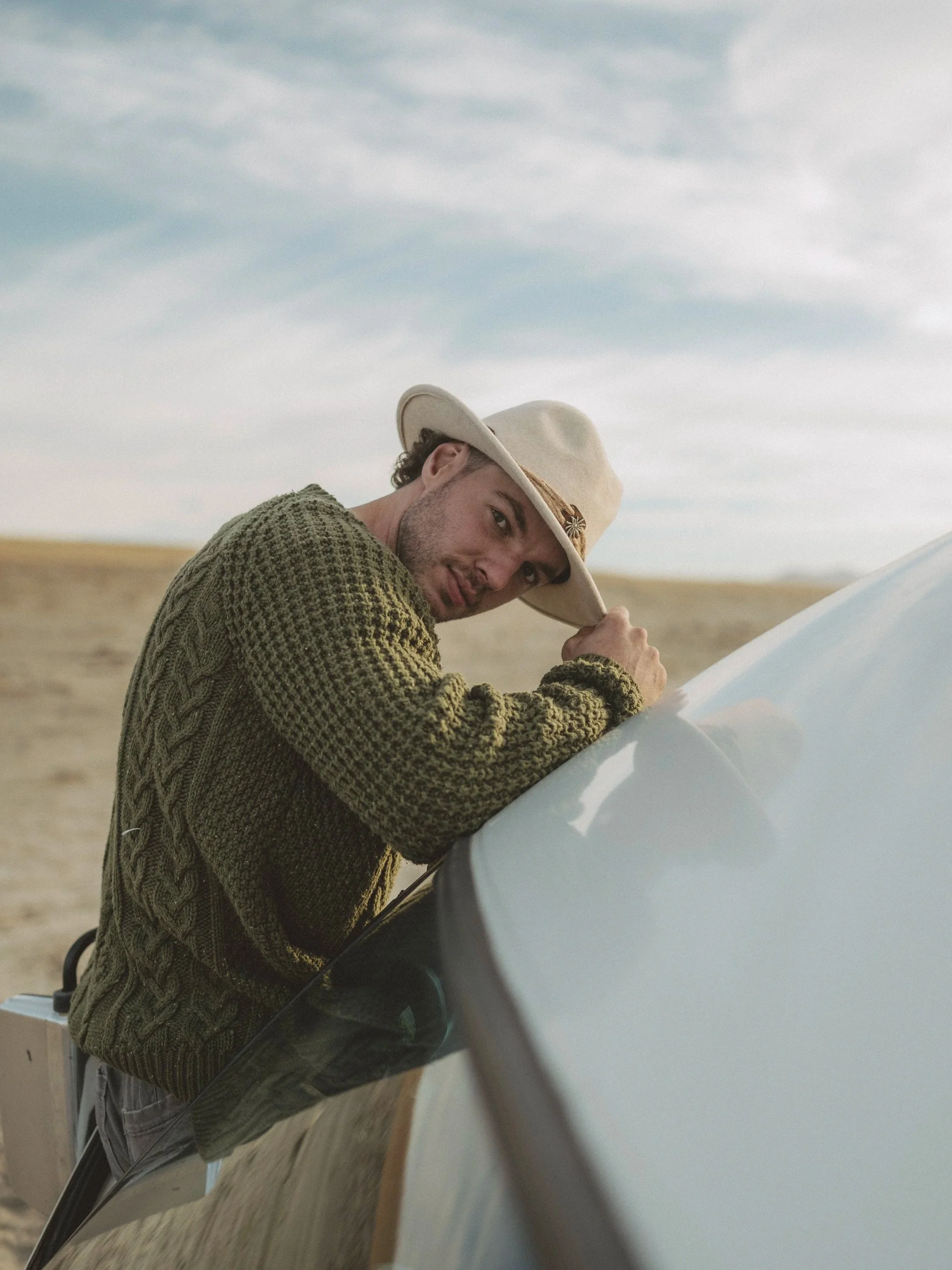 A man with curly hair and a beard, wearing a wide-brimmed hat and a green knit sweater, leaning on the hood of a car in an open, grassy field under a cloudy sky.