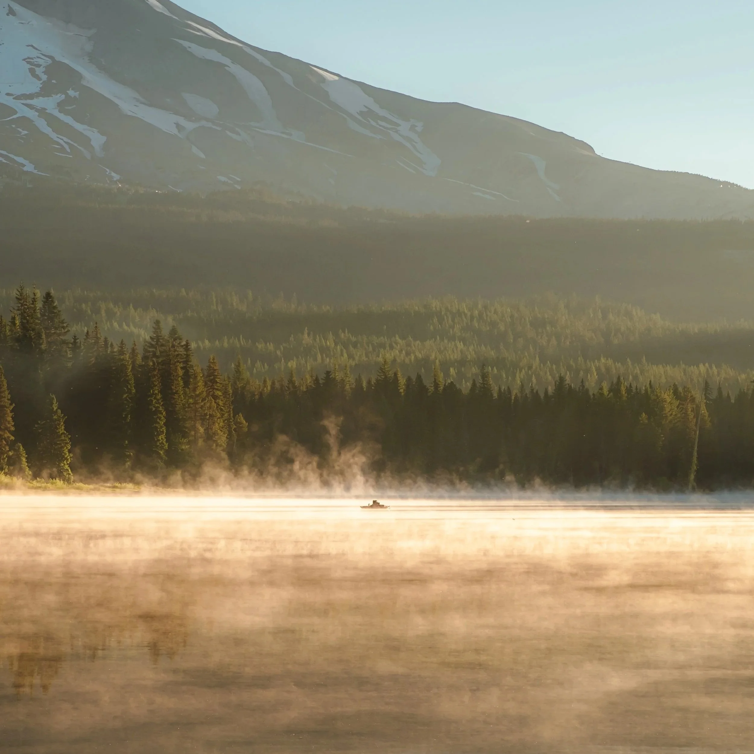 A boat on a foggy mountain lake at sunrise with a backdrop of forest and snow-capped peaks.