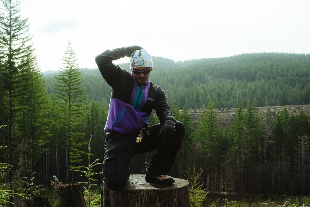 A person dressed in outdoor gear, including a hat, sunglasses, and gloves, poses with one foot on a tree stump in a forest with tall green trees and mountains in the background.