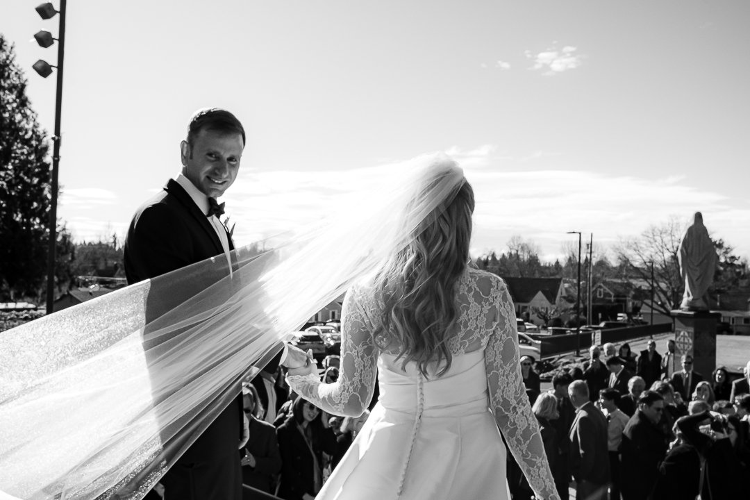 Bride with long, wavy hair in a lace wedding dress and veil, holding the groom's hand, smiling at him during an outdoor wedding ceremony. Groom in a tuxedo is smiling and looking at the bride, with guests and a statue in the background.
