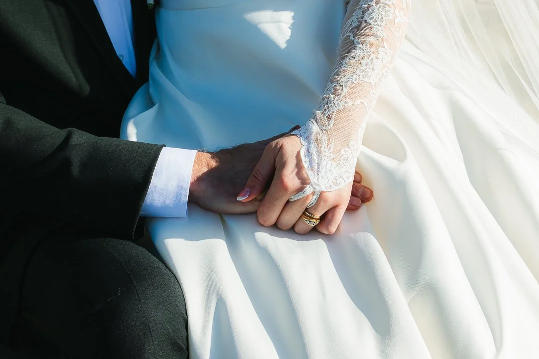 Close-up of a groom and bride holding hands, with the bride's hand wearing a lace sleeve and wedding ring, and the groom's hand in a suit, on a white wedding dress fabric.