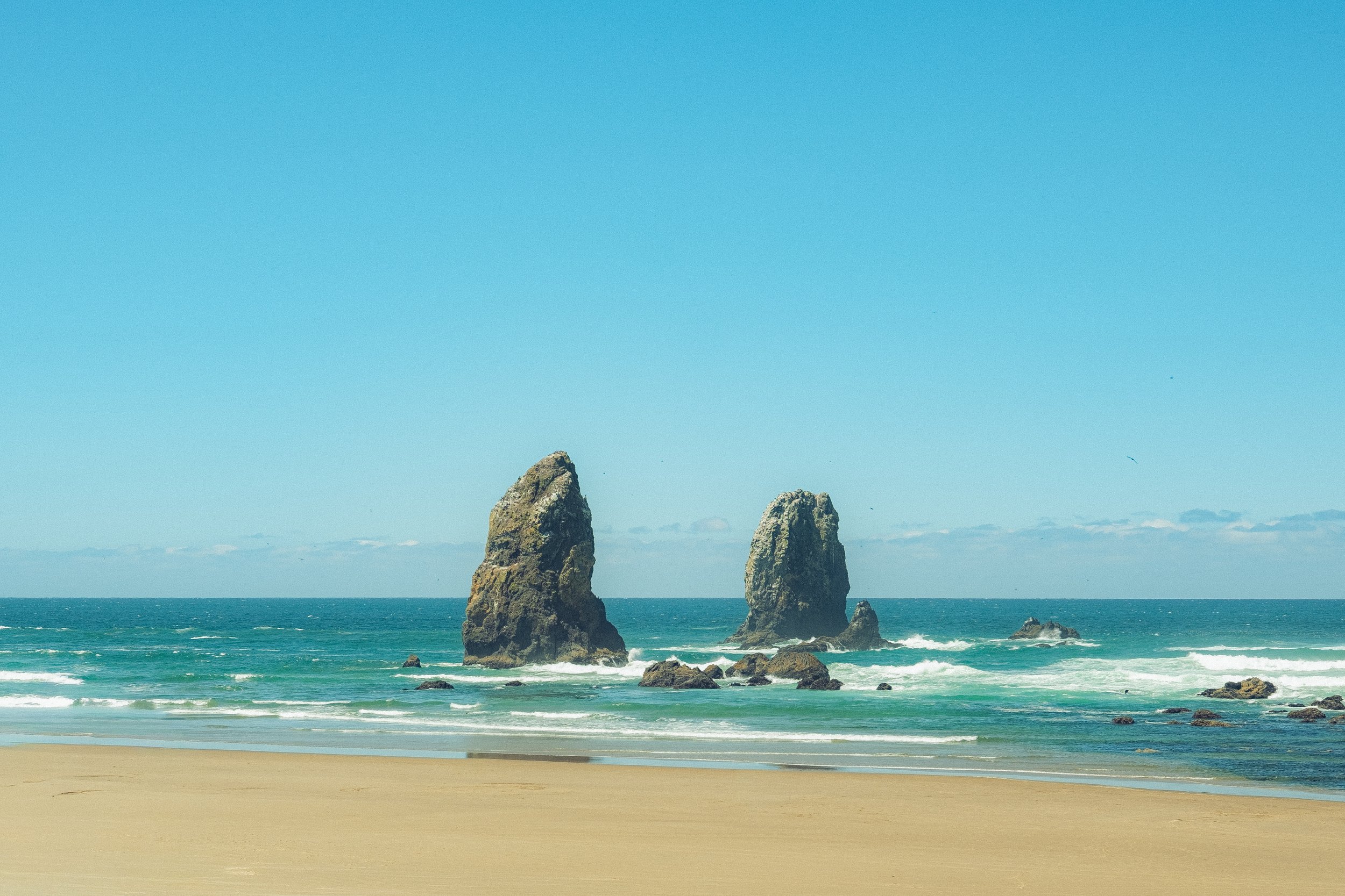 A beach with light-colored sand, turquoise water, and three large rock formations rising from the ocean under a clear blue sky.