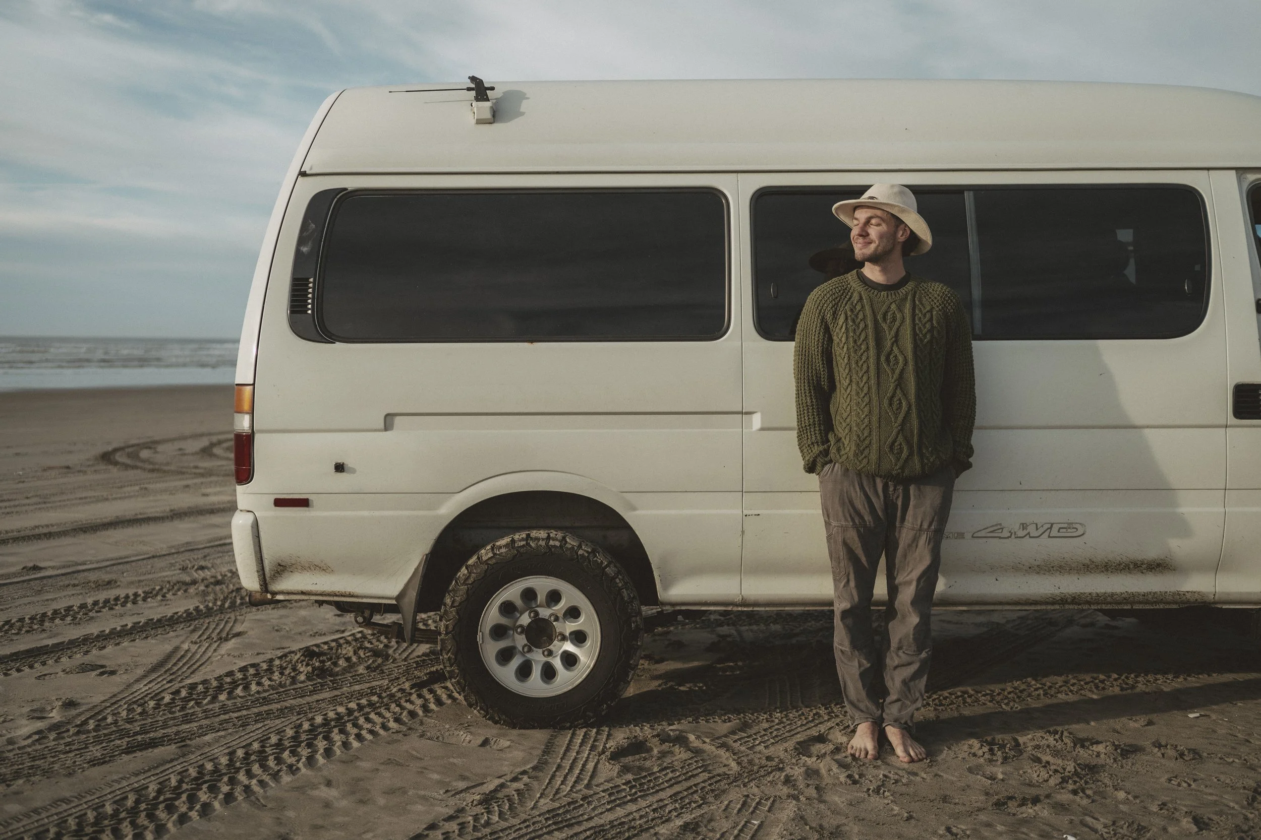 A man standing barefoot on a sandy beach in front of a white van, wearing a wide-brimmed hat and a green sweater, with the ocean and sky in the background.