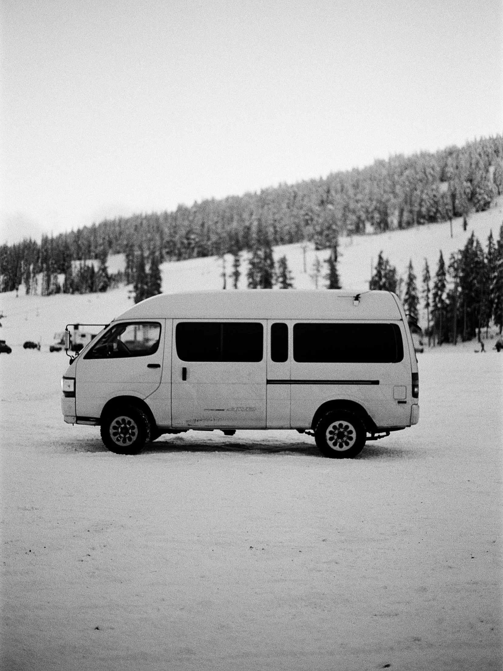 A white van parked on snow-covered ground with a background of snow-covered hills and tall pine trees in a winter landscape.