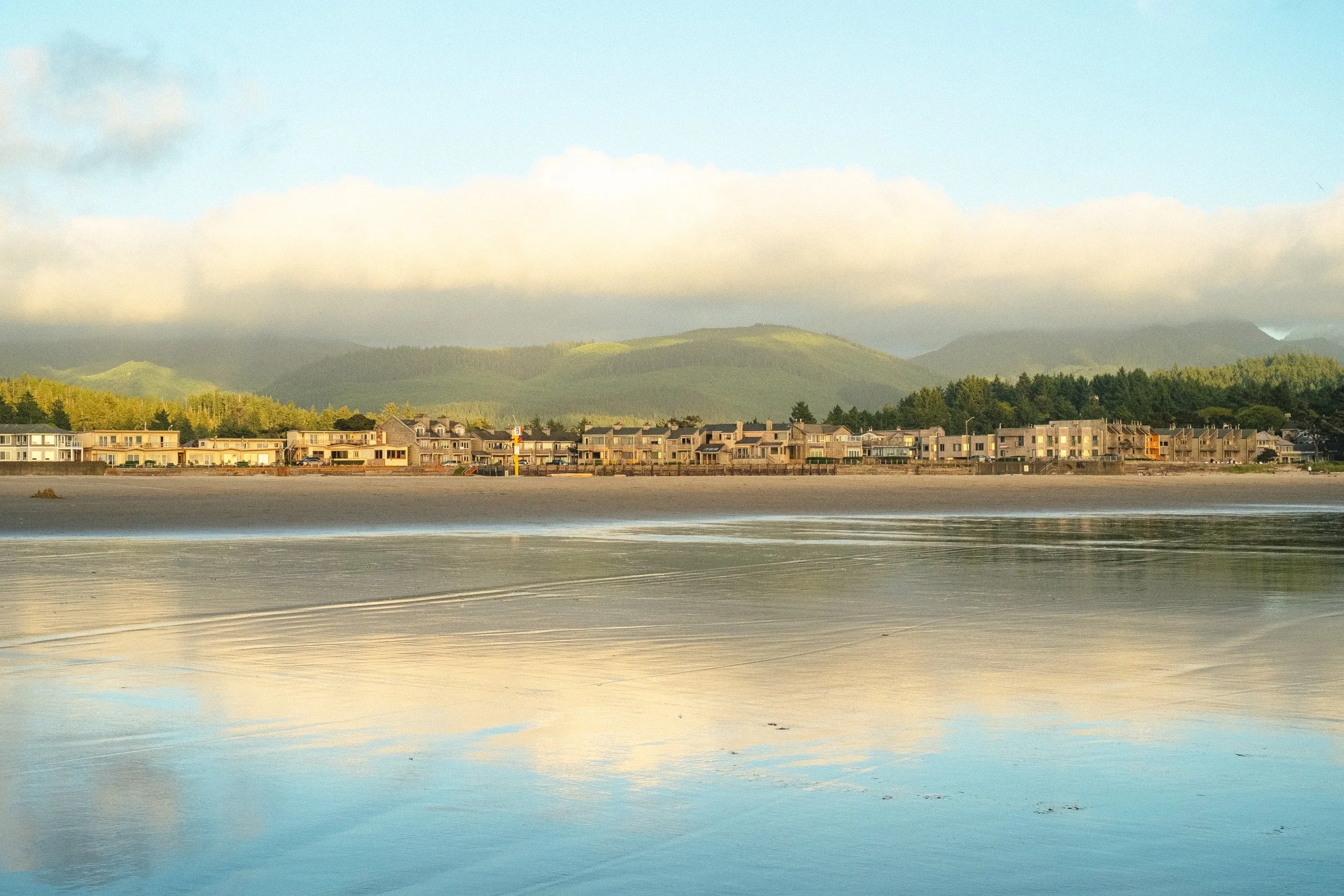 View of a row of houses along the water with green hills and mountains in the background under a cloudy sky.