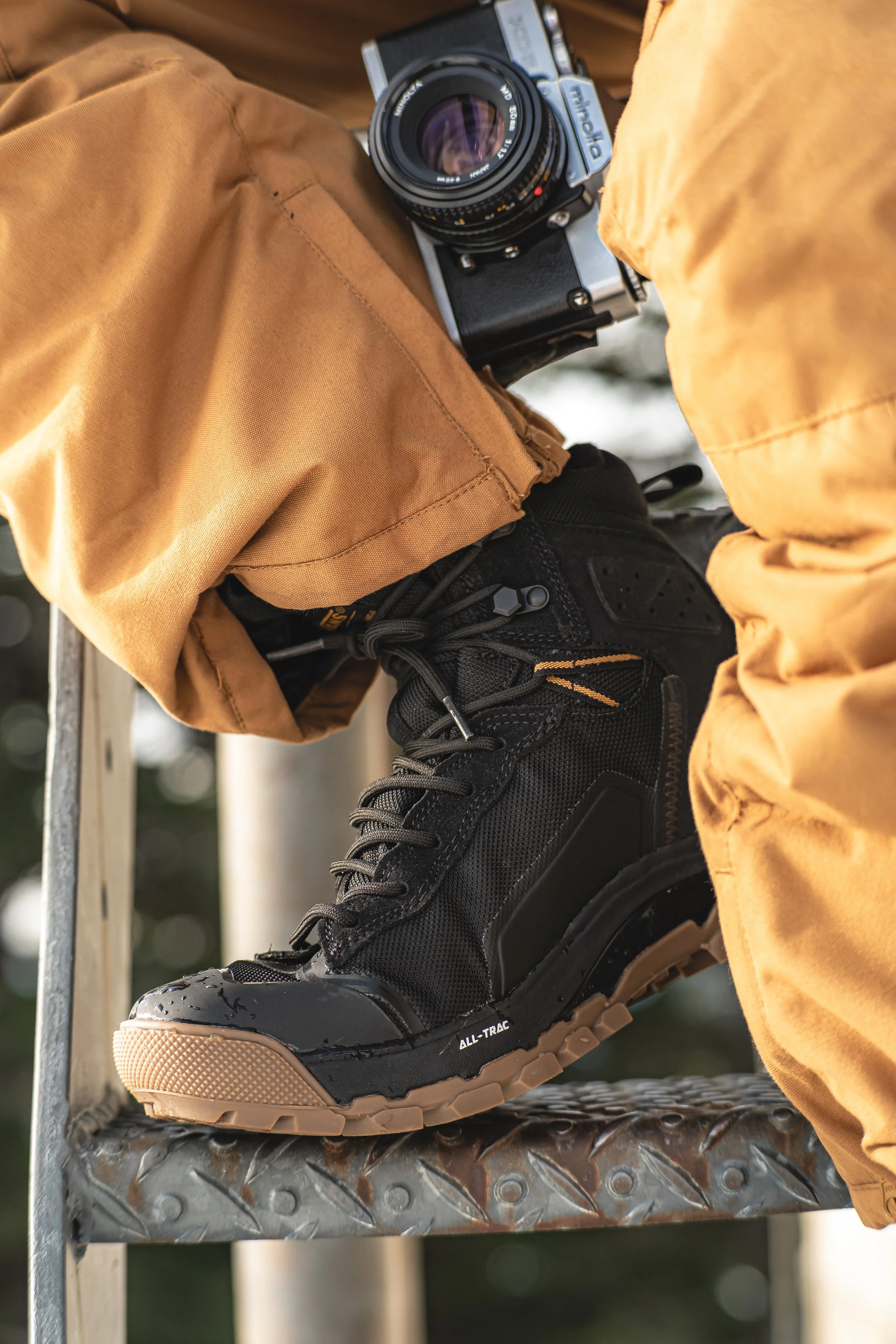 Close-up of a person's black hiking boot on a metal ladder step, with a brown pant cuff and a film camera resting on the knee.