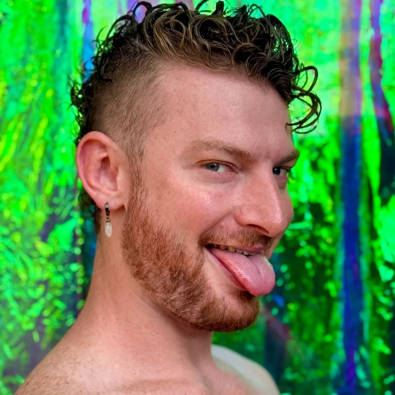 A young man with curly hair and a beard sticking out his tongue, wearing a dangling crystal earring, against a colorful, iridescent background.