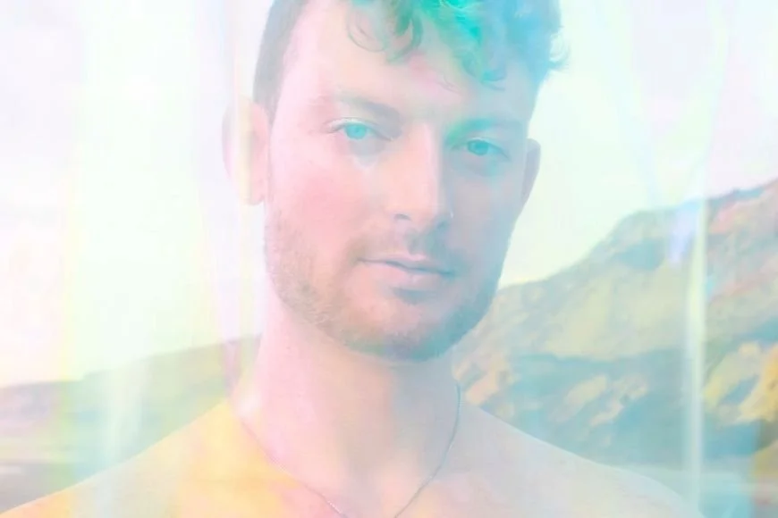 A person with short curly hair and facial hair, looking towards the camera, with a colorful, blurred background.