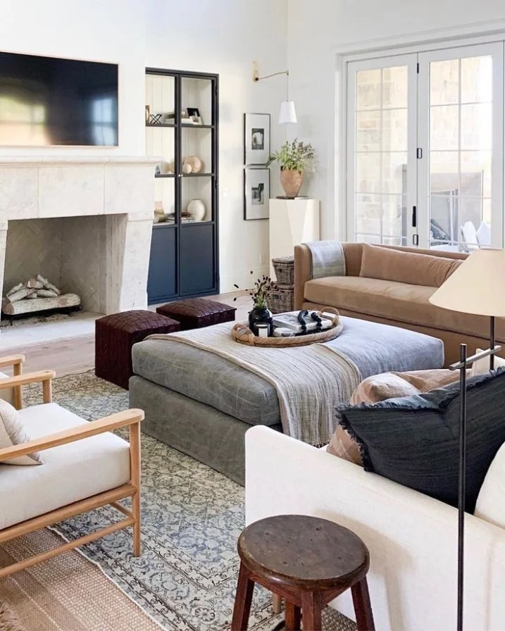 A cozy living room with a light-colored fireplace, a large flat-screen TV, a black display cabinet, framed pictures, a potted plant, a sliding glass door, and various seating including a beige sofa, a white armchair, and a tan chaise lounge. There is a magazine tray on an ottoman, a small wooden stool, and decorative pillows.