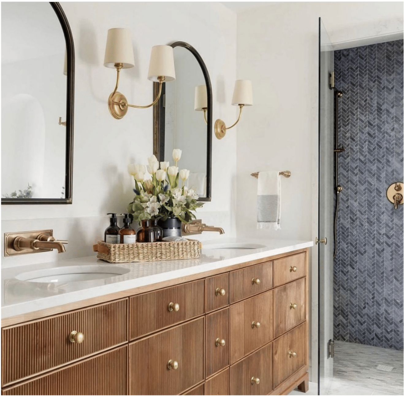 Bathroom with a double sink vanity, large mirrors, brass fixtures, wall sconces, and a shower with dark tile and a glass door.