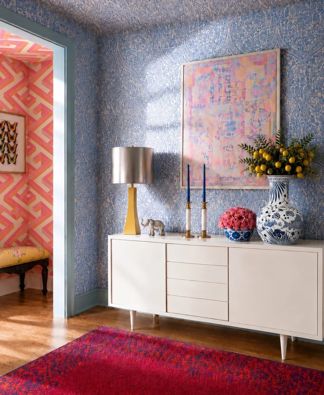Decorated interior with a white sideboard, blue patterned wallpaper, colorful abstract painting, yellow and blue vases with flowers, two blue candles, a table lamp with yellow base and silver shade, and a red rug. An adjoining room has pink geometric wallpaper, butterfly artwork, and a yellow upholstered bench.