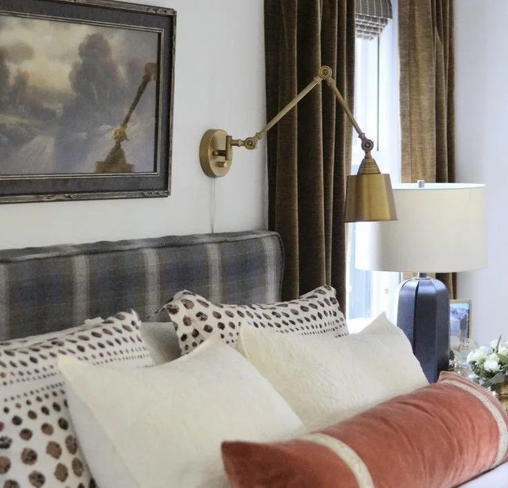 A cozy bedroom with a plaid headboard, patterned pillows, and a brass wall lamp.