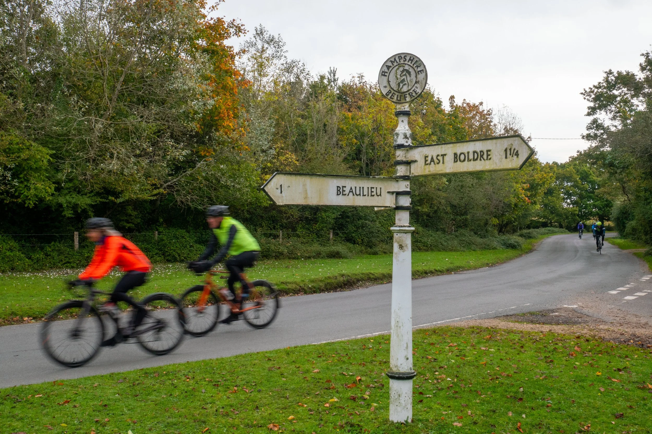 Cyclists riding bikes along a curved road, with a sign indicating directions to East Boldre and Beaulieu, surrounded by trees with fall foliage.